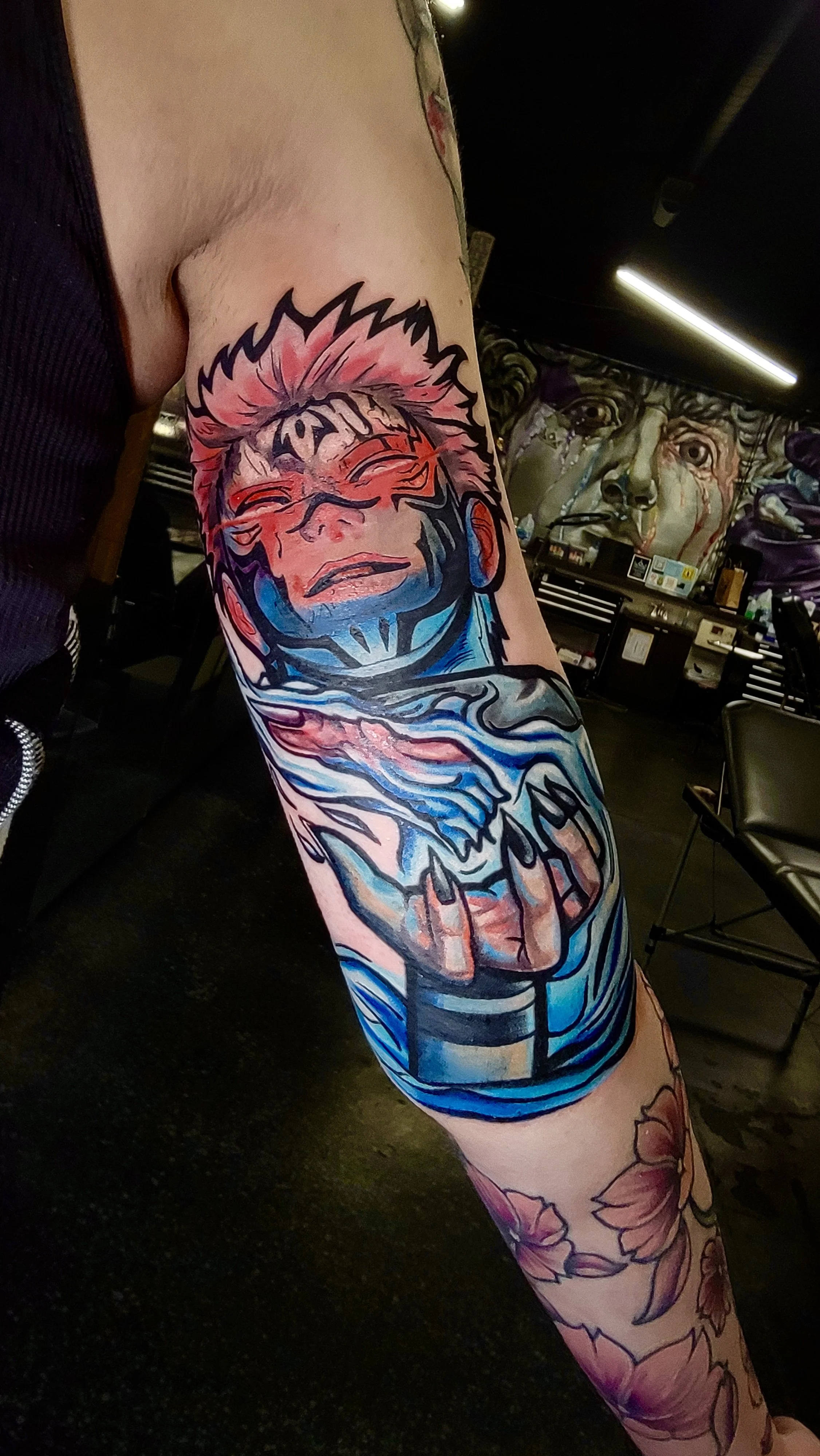 Colorful tattoo sleeve featuring a person with spiky hair, tears, and a hand holding a bone on a person's arm. In the background, a mural of a face with expressive eyes and dripping tears is visible.