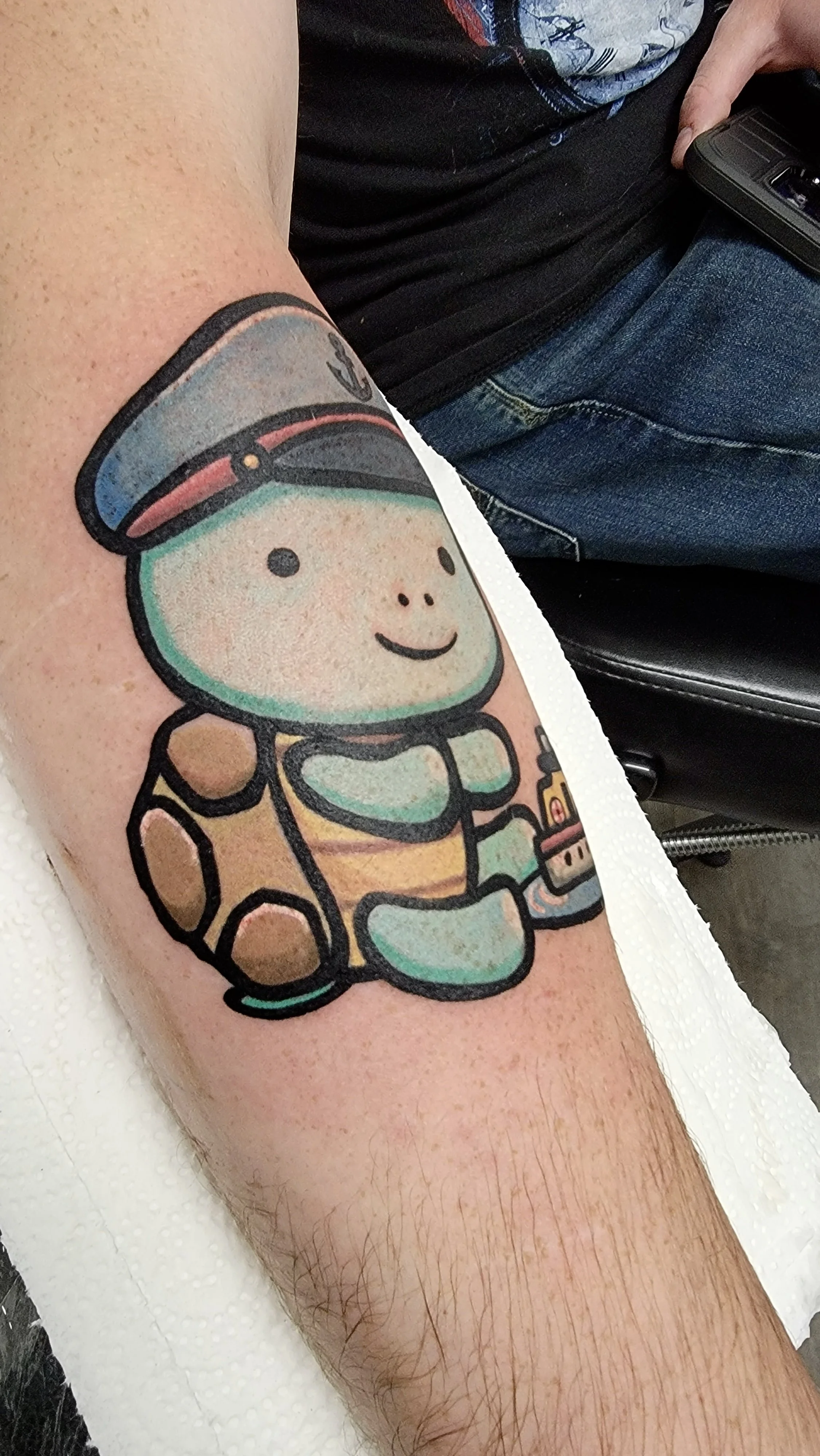 A colorful tattoo of a smiling cartoon astronaut girl is on a person's arm. The astronaut has a round face, a space helmet, and a spacesuit with gloves, sitting on a small planet or moon.