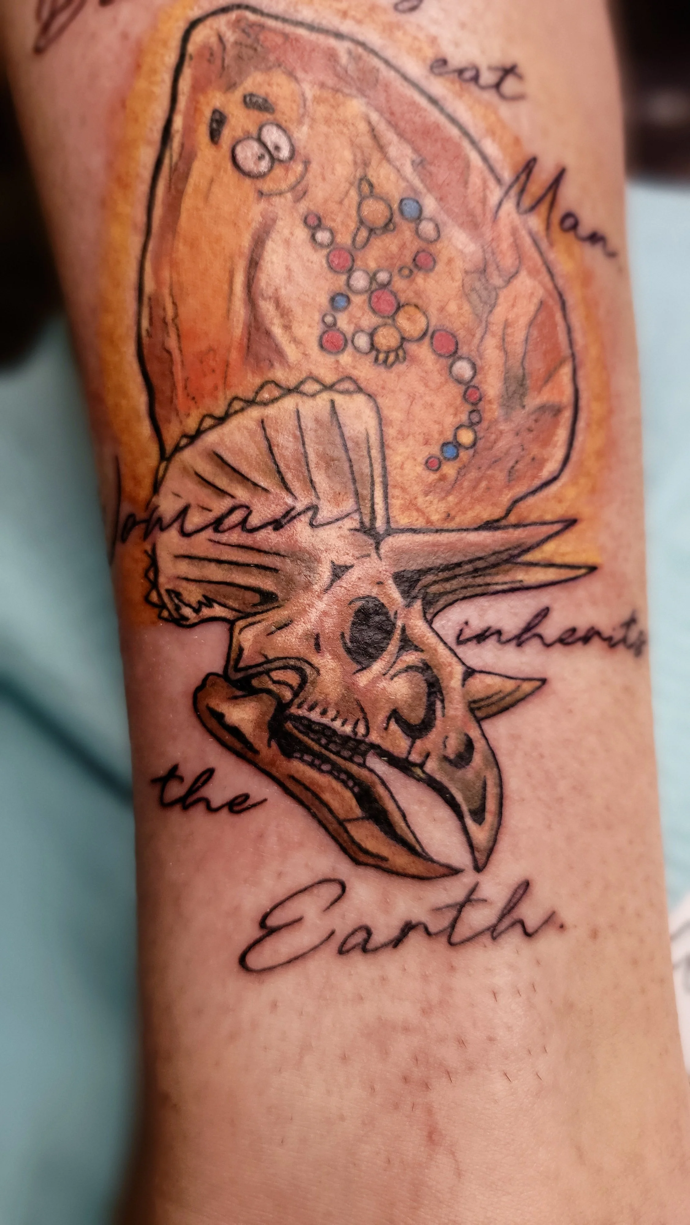 A tattoo of a detailed ram's skull with curved horns, overlaid with a colorful abstract pattern and surrounded by cursive text.
