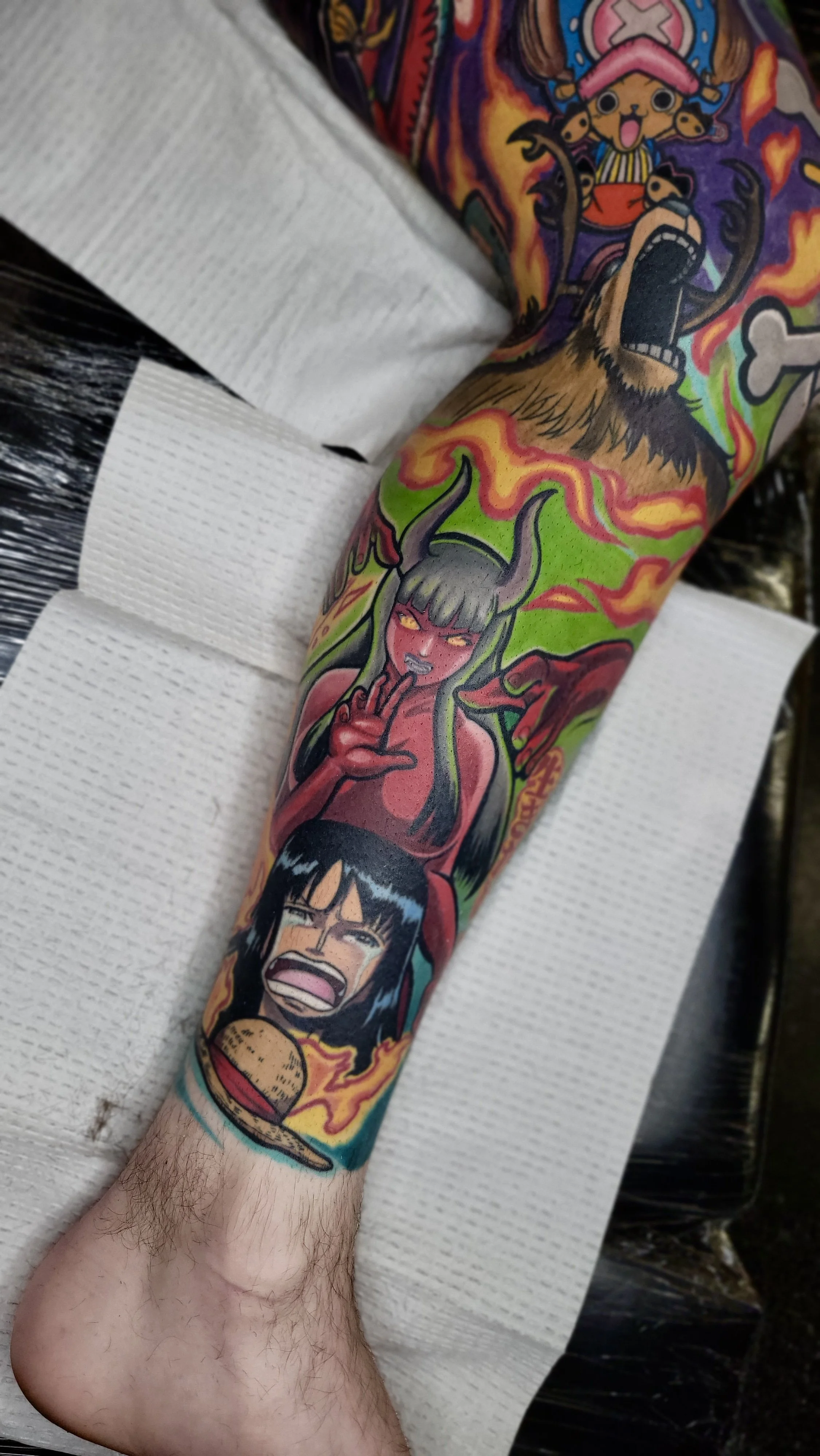 Colorful tattoo sleeve featuring a female demon with green hair, horns, and red skin, holding a cigarette, with other characters, flames, and surreal elements.