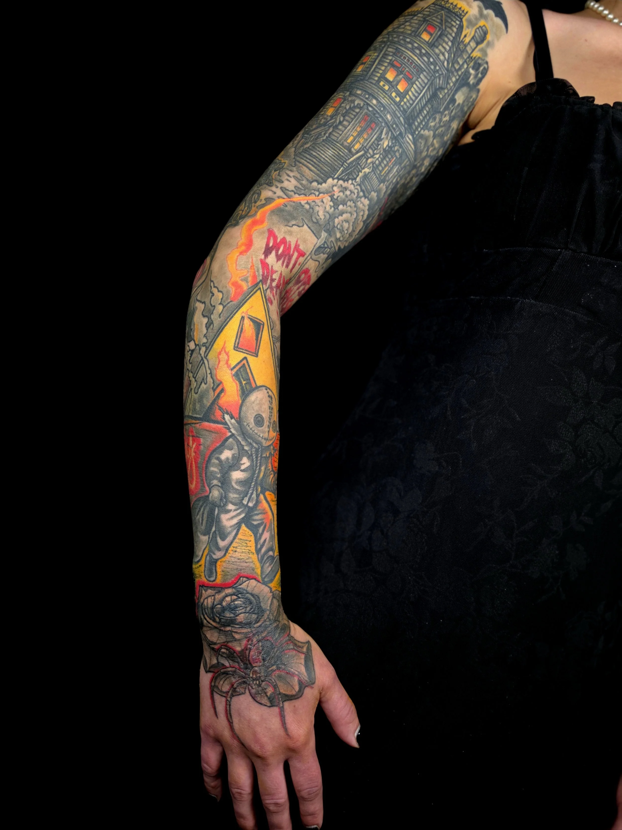 A person's arm with colorful tattoos of a building, smoke, flames, a skull, and a spider. The person is wearing a black sleeveless top and black pants, with a pearl bracelet visible on the wrist.