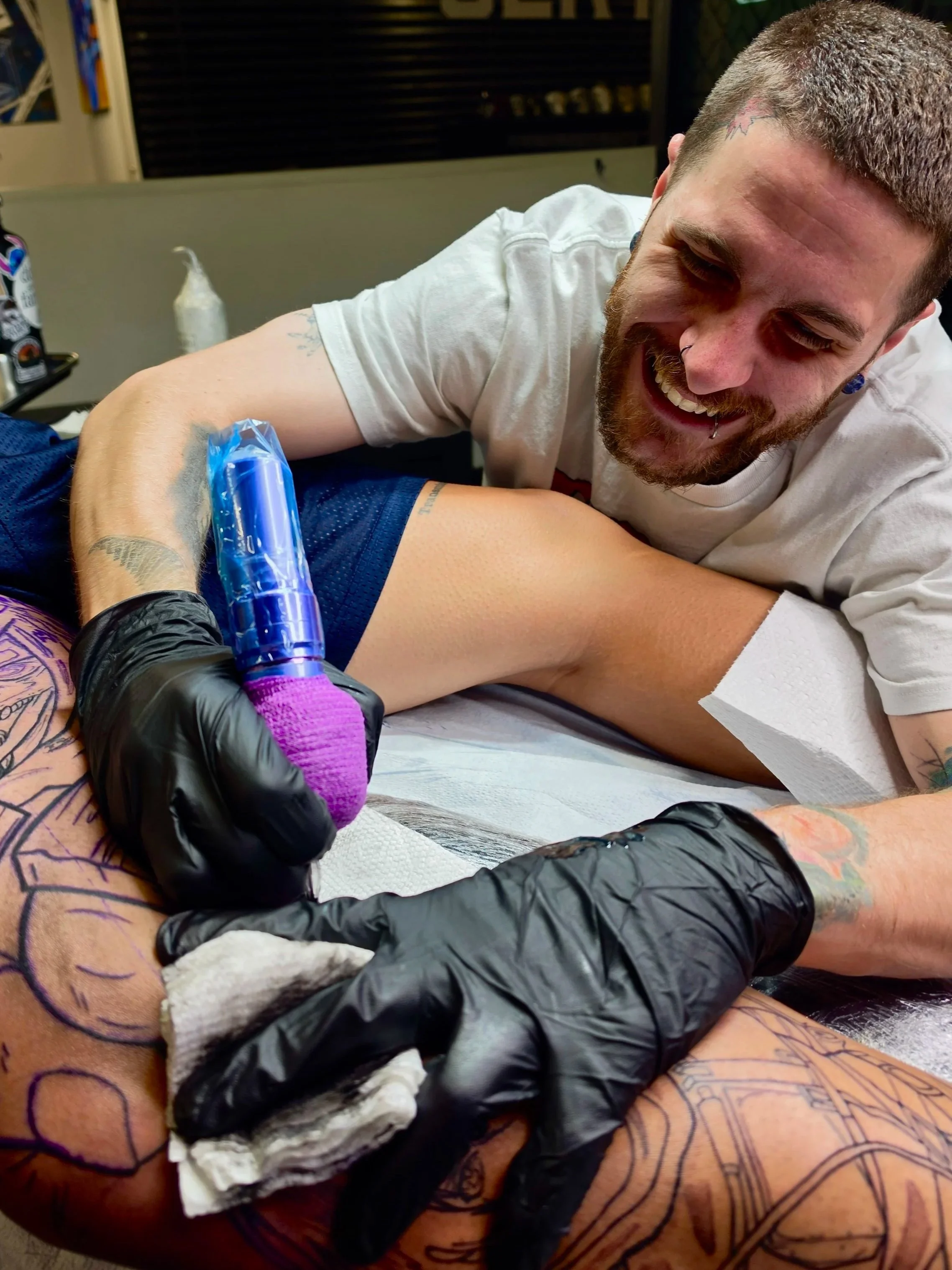 A man with tattoos and a beard joyfully getting a tattoo on his thigh from a tattoo artist wearing black gloves.