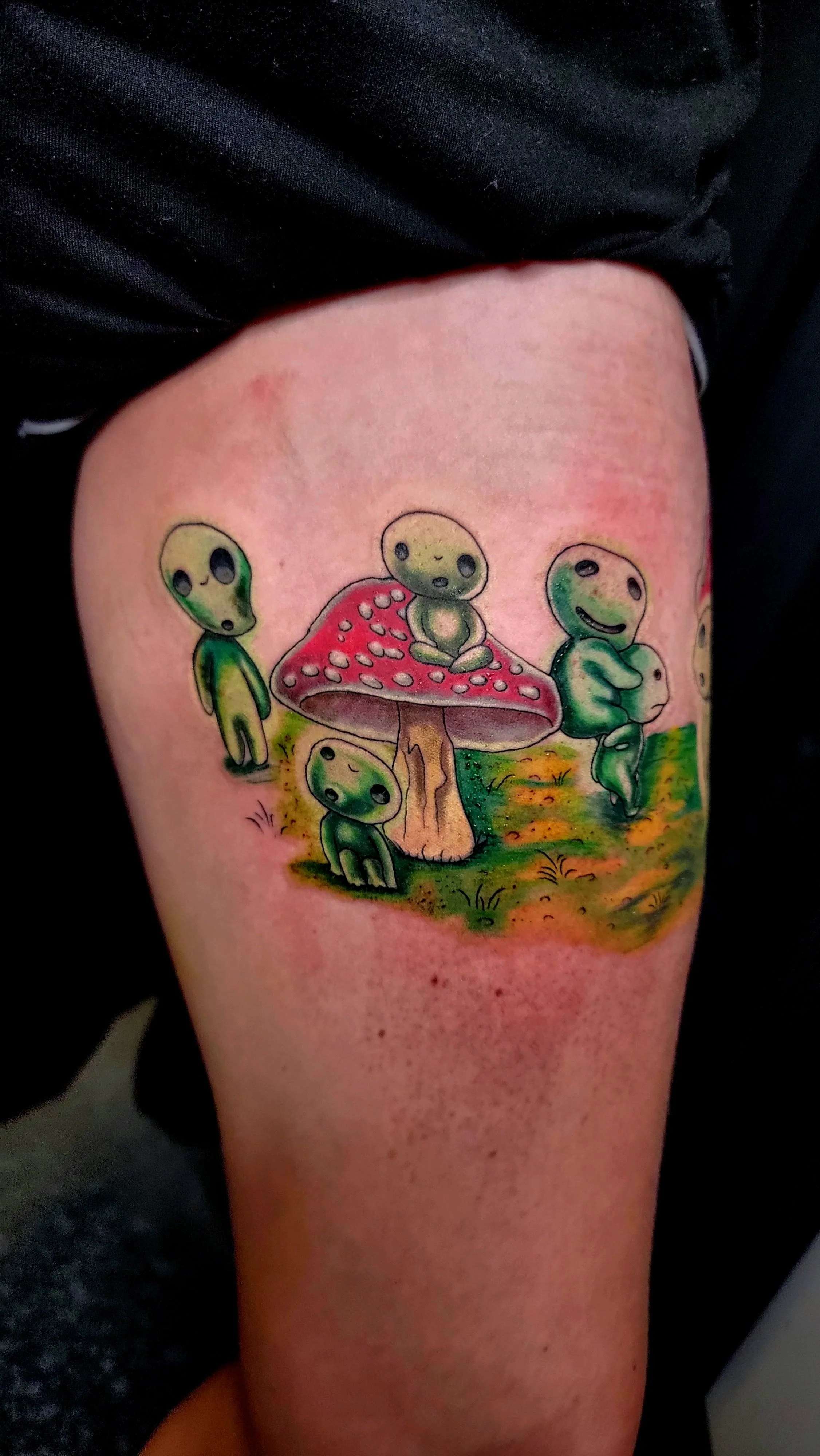 Colorful tattoo of cute green alien characters around a red mushroom with white spots, set in a whimsical outdoor scene.