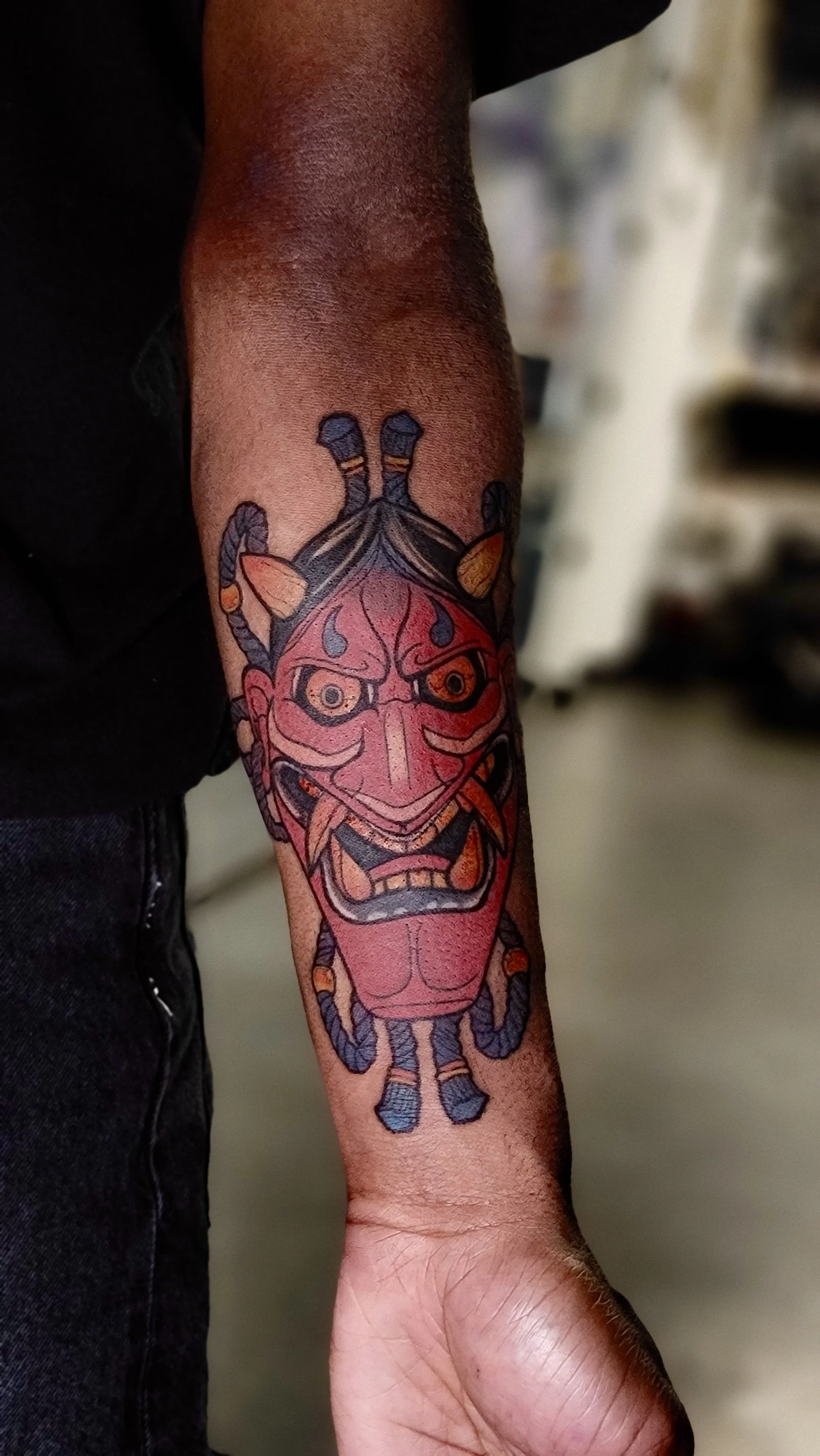 Tattoo of a traditional Japanese demon mask on a person's forearm, with a red face, large angry eyes, and blue and black hair.