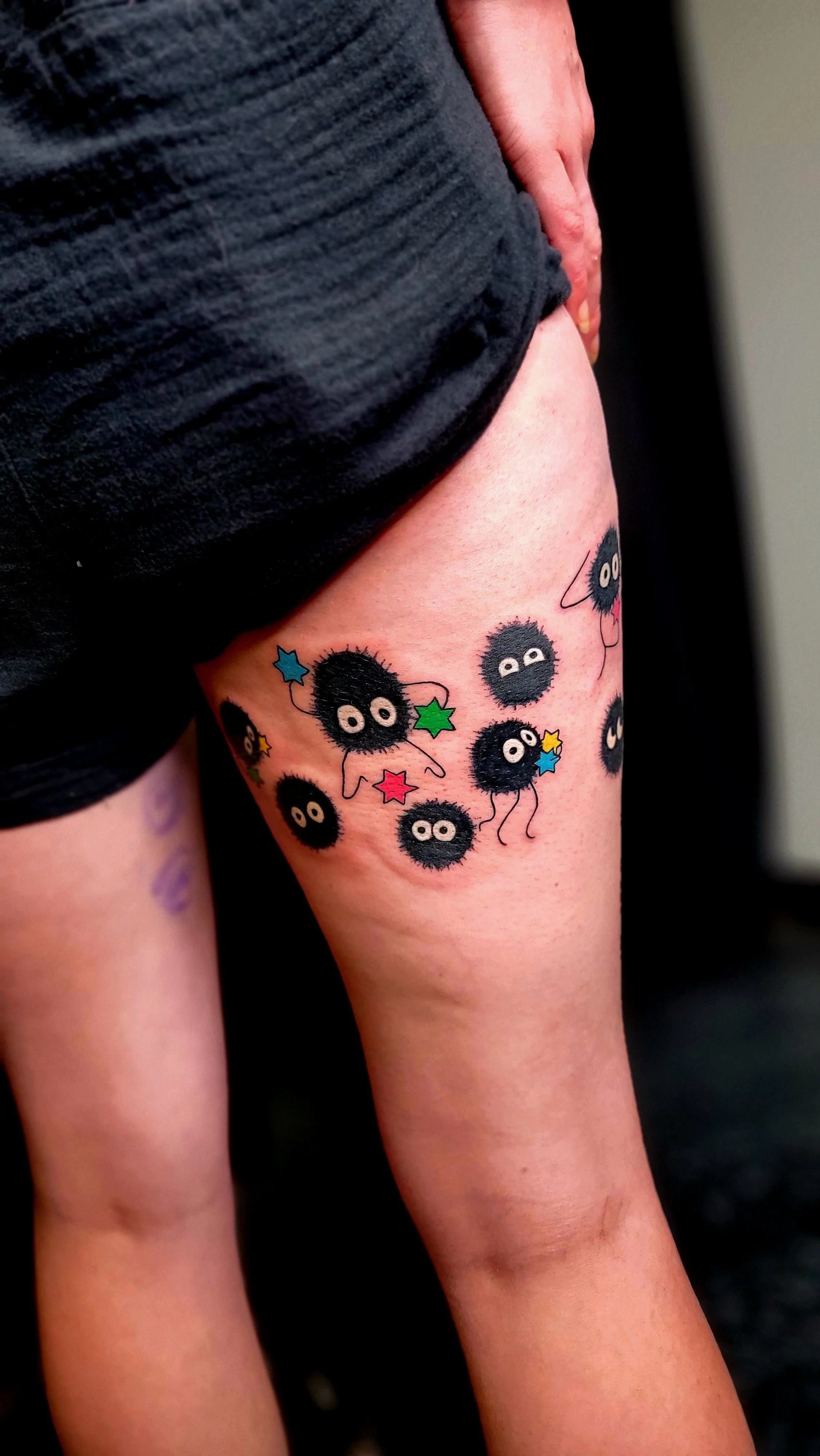 A person's upper thigh showing a colorful tattoo featuring multiple small black fuzzy creatures with big white eyes, surrounded by multicolored stars.