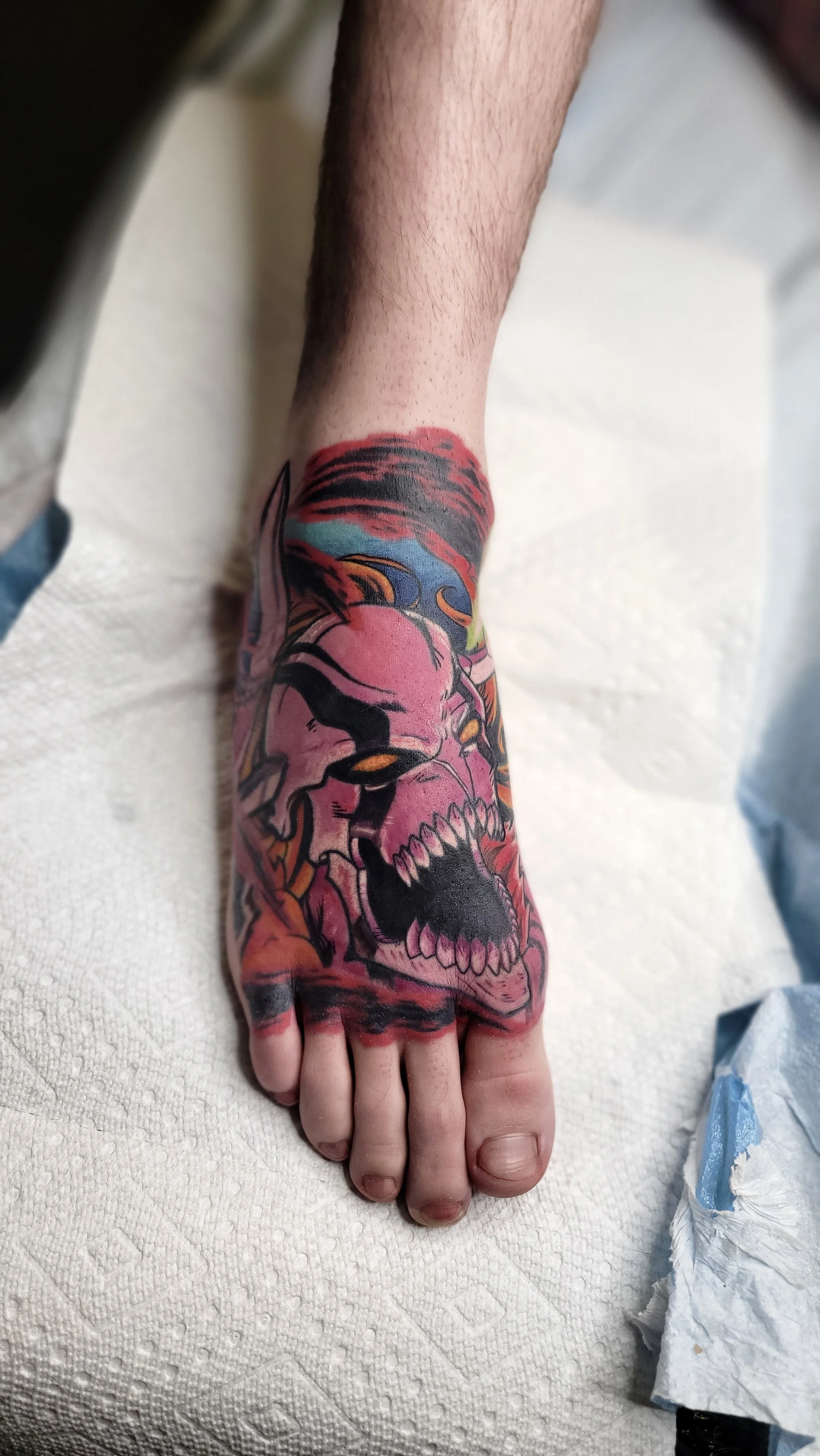 Colorful tattoo of a skull on a person's forearm with a vibrant background, resting on a white textured surface.