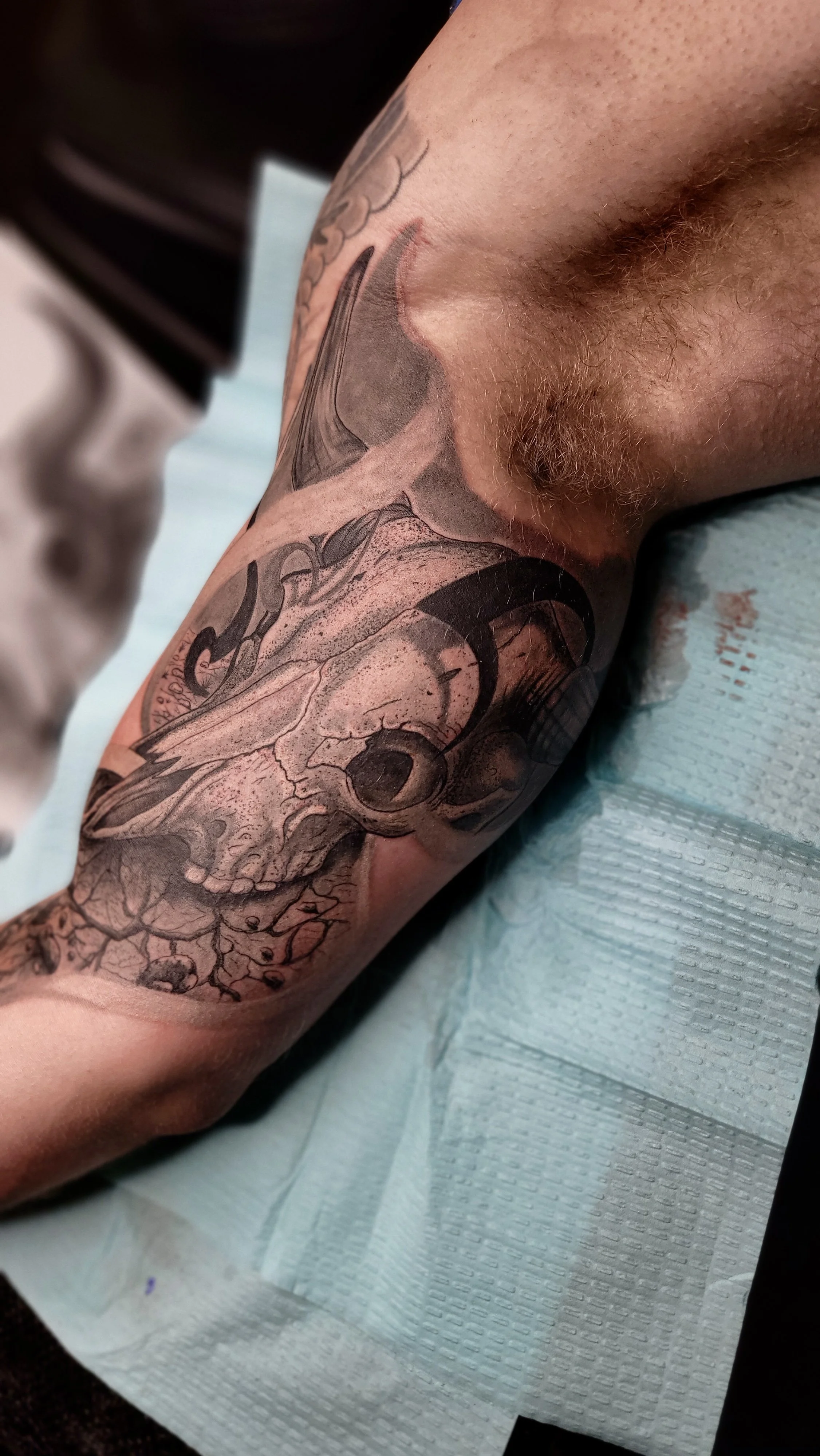 Close-up view of a person's arm with a detailed black and gray tattoo of a skull with cracks and an eye socket, surrounded by intricate linework and shading.