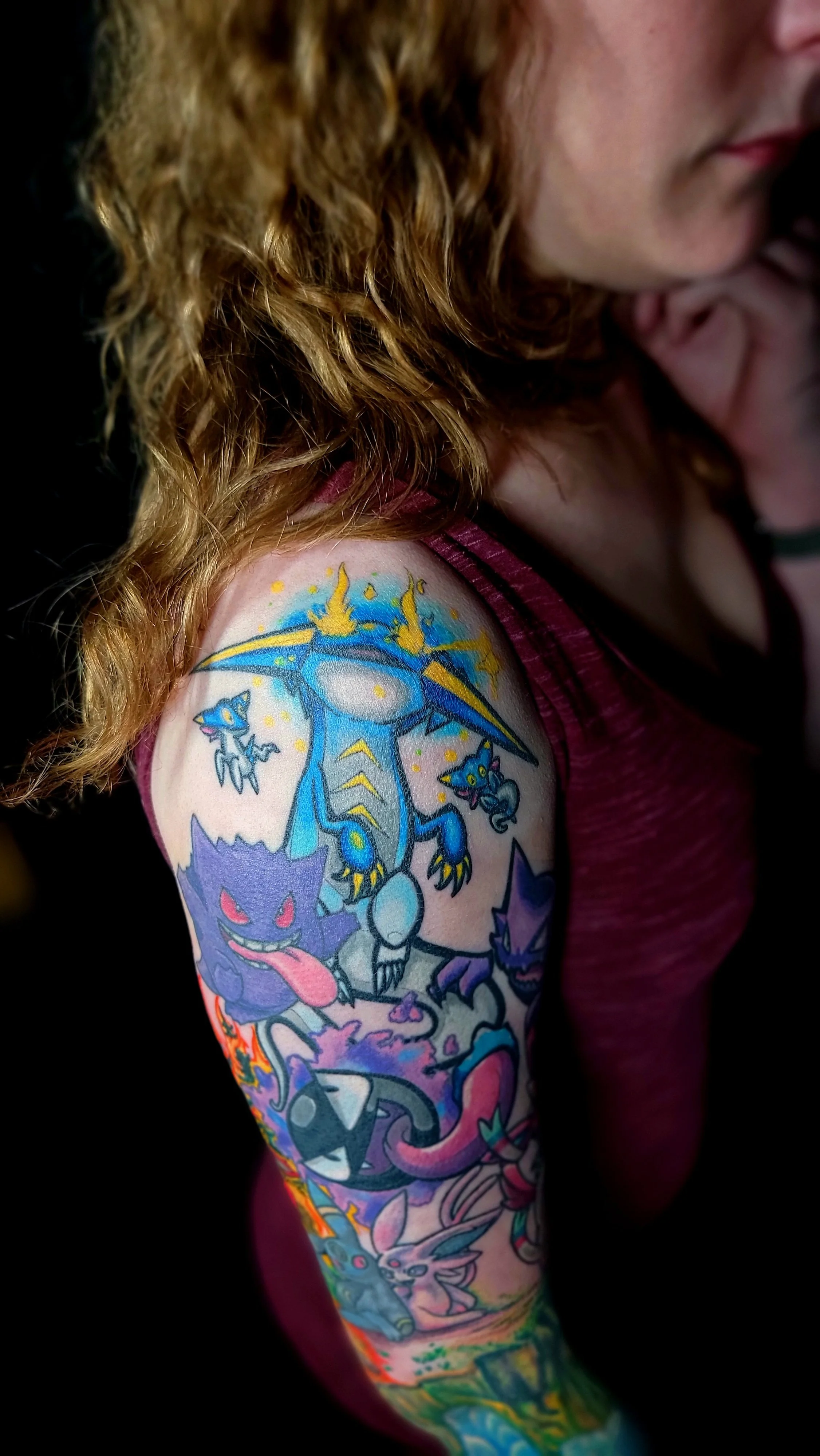 A person's arm with a colorful tattoo sleeve featuring various fantasy and cartoon characters, including dragons, cats, and other creatures.