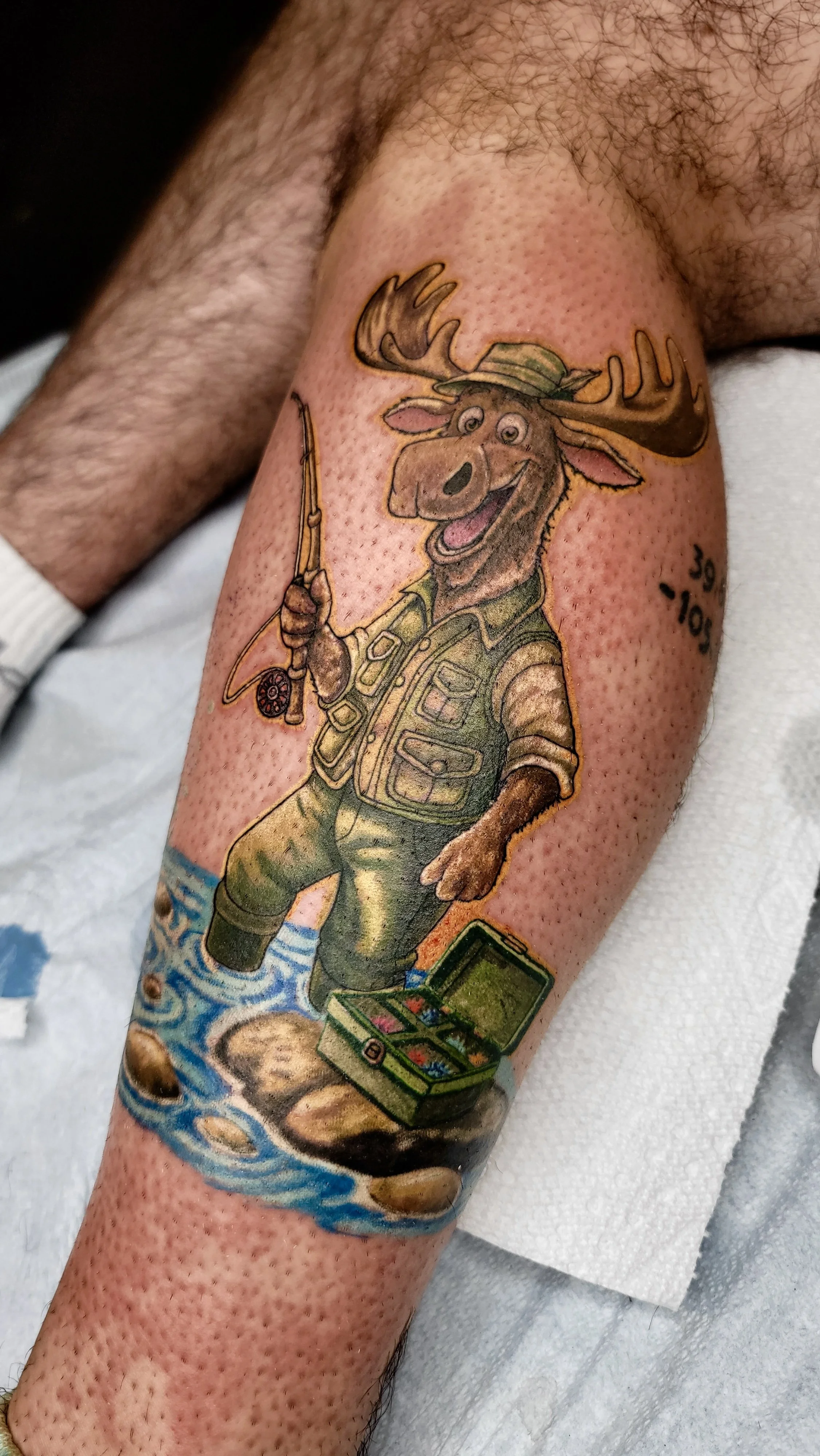 Colorful tattoo of a moose dressed as a fisherman, holding a fishing rod and standing in water with rocks. There is a fishing tackle box at the bottom.
