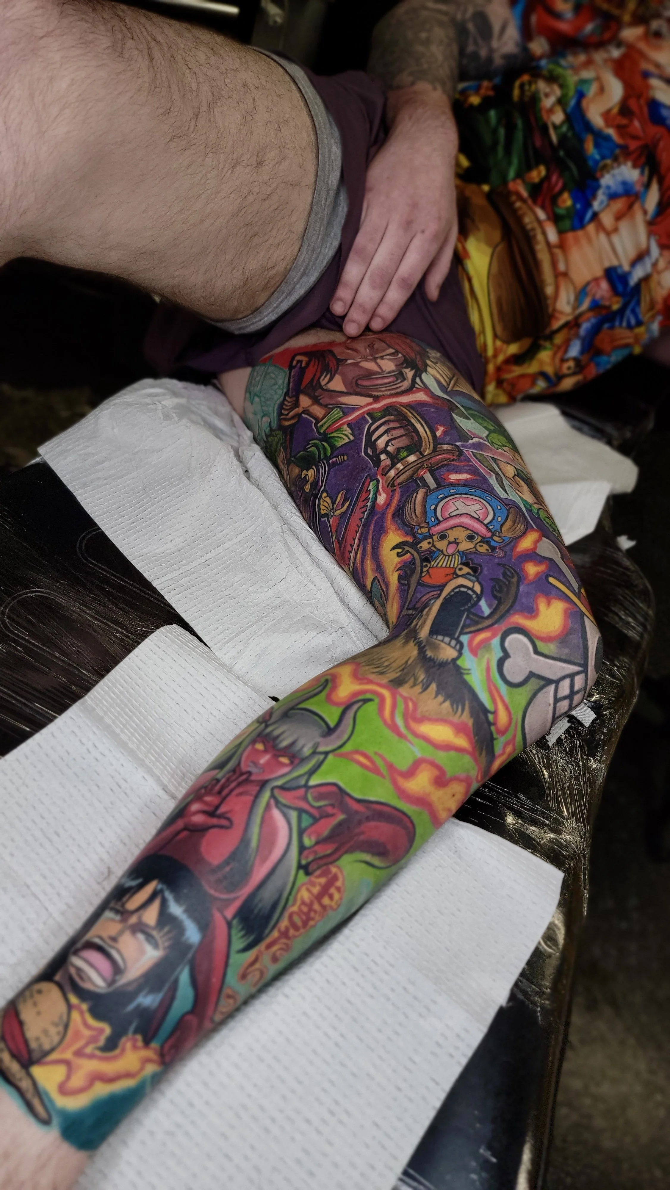 A person's arm with a colorful tattoo sleeve featuring characters from a video game, lying on a white paper towel on a tattooing surface.
