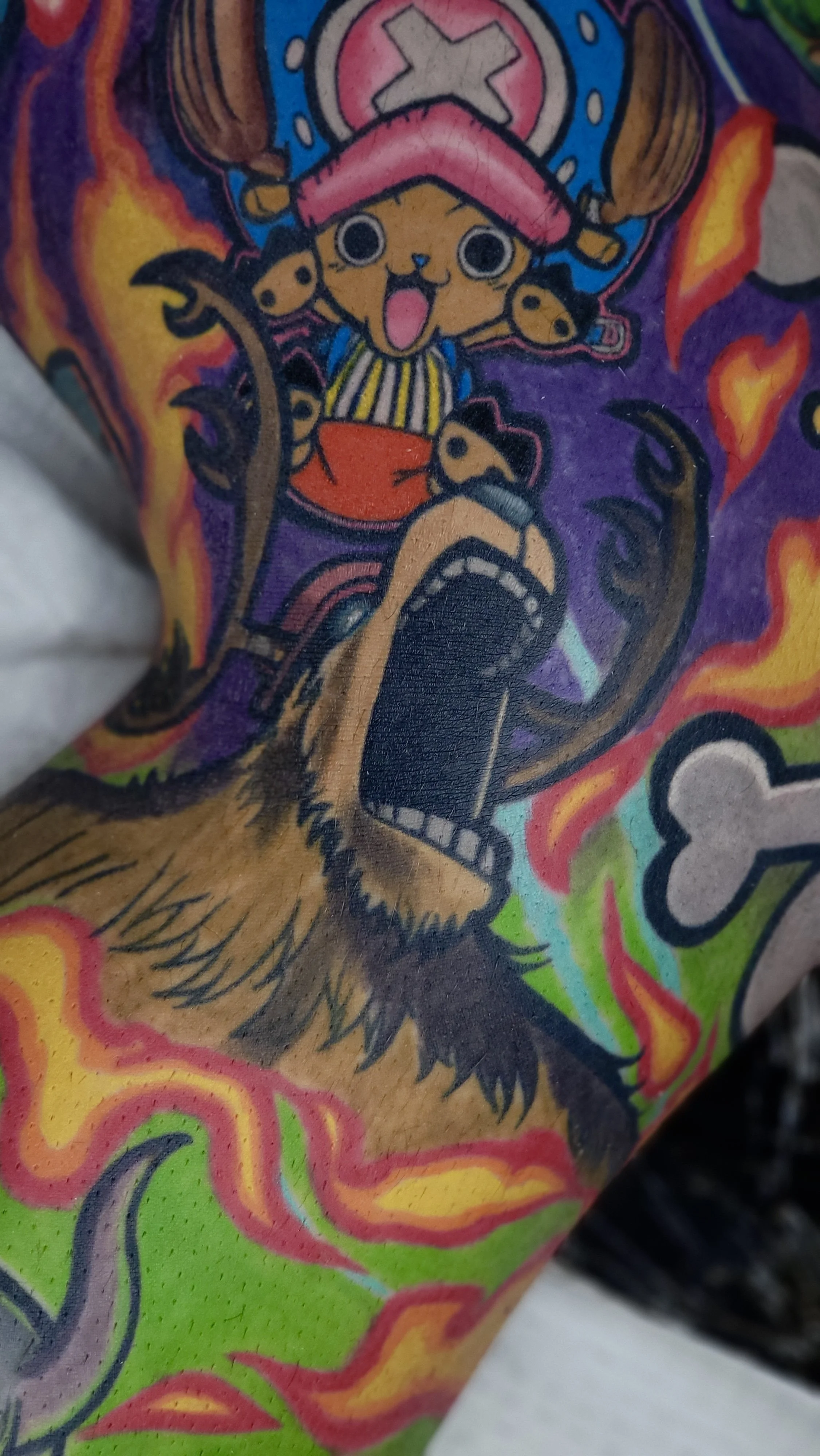 Colorful tattoo of a cartoonish character resembling Luffy from One Piece, wearing a pink hat with an X and a striped shirt, surrounded by flames and vibrant colors.