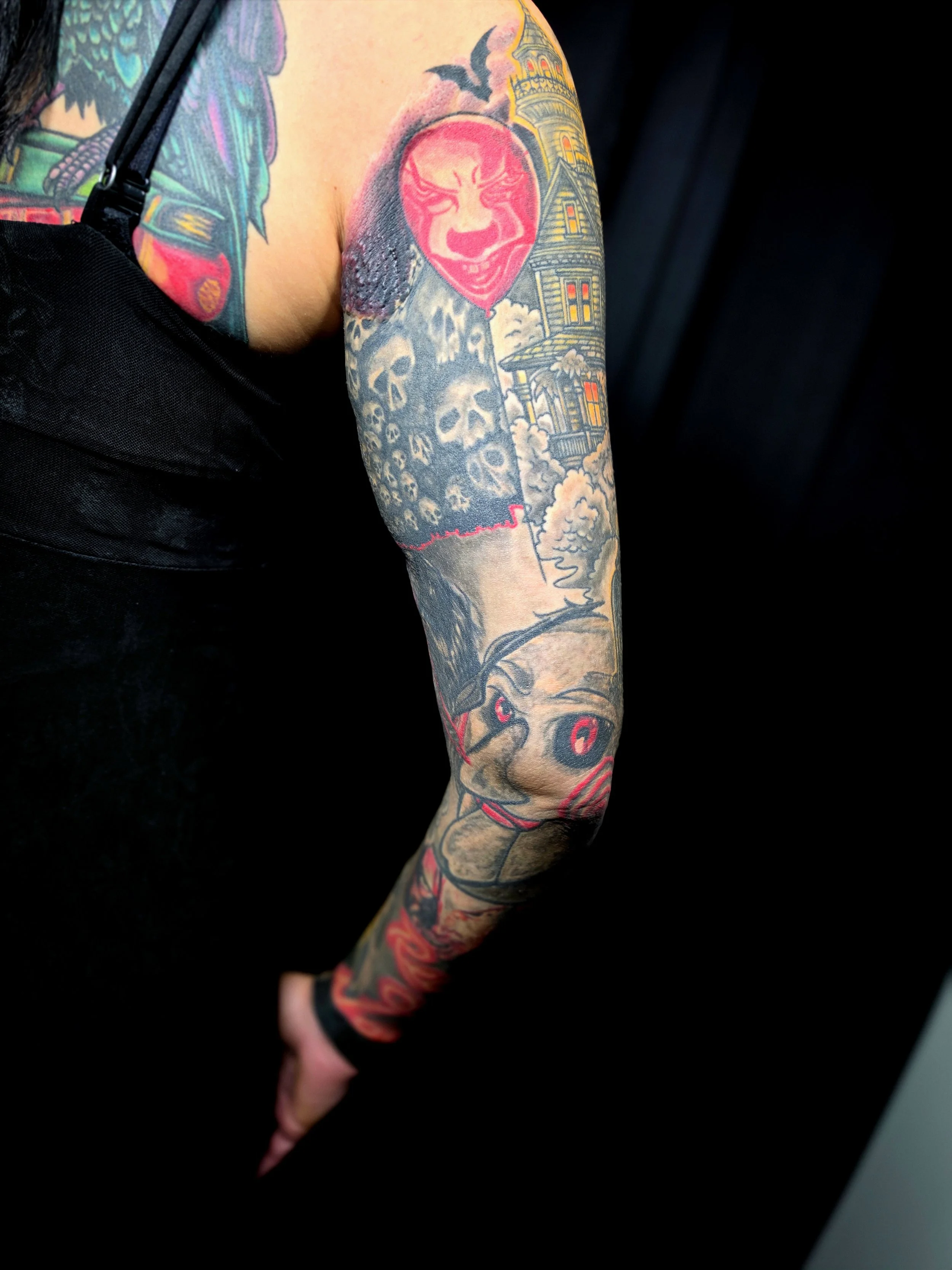 Close-up of a person's tattooed arm featuring various designs, including a red face with snarling expression, skulls, clouds, and a detailed building or castle.
