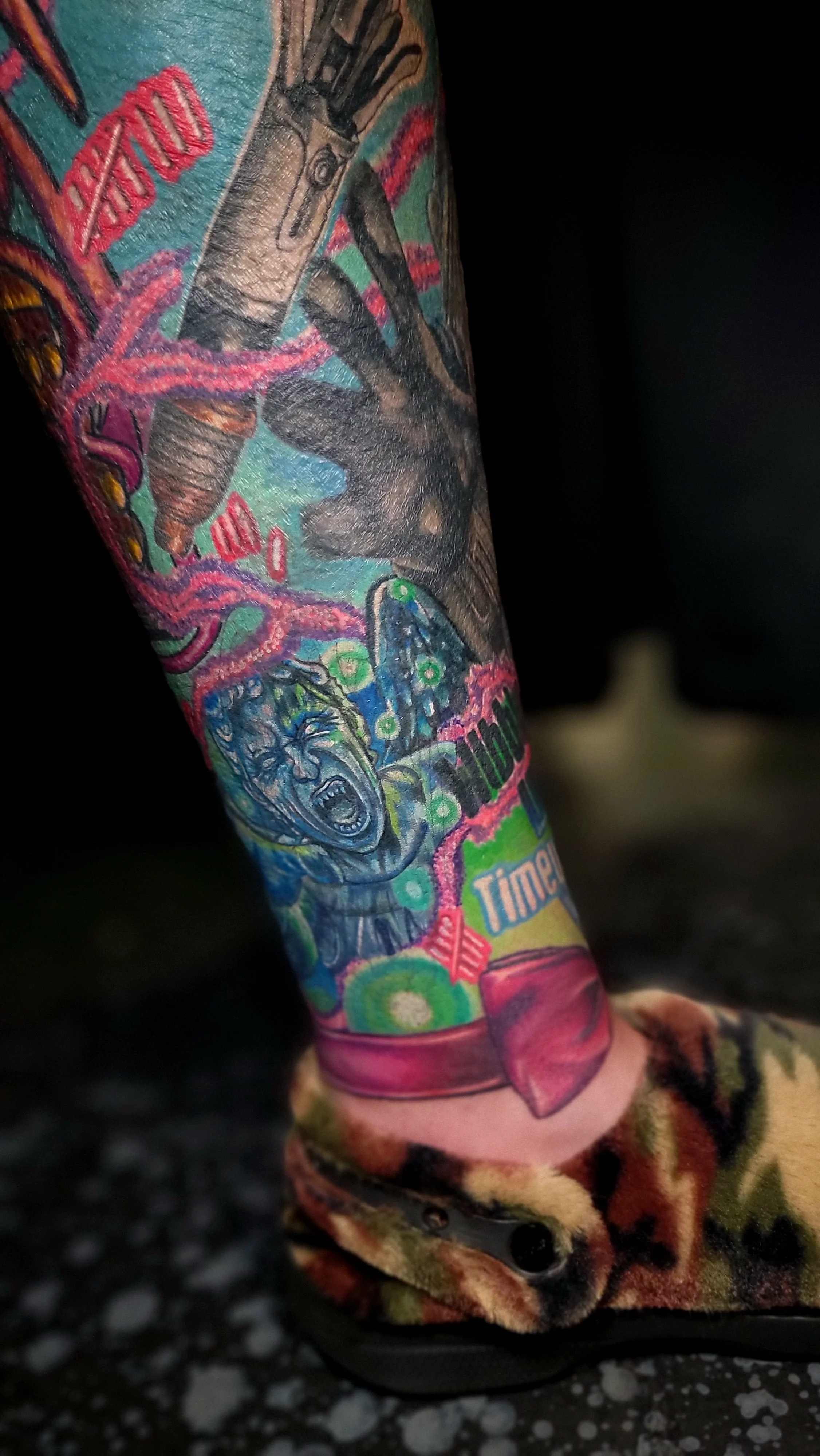 Colorful tattoo on a person's arm featuring a hand holding a mechanical object, a crying face, and various abstract and graffiti-style designs.