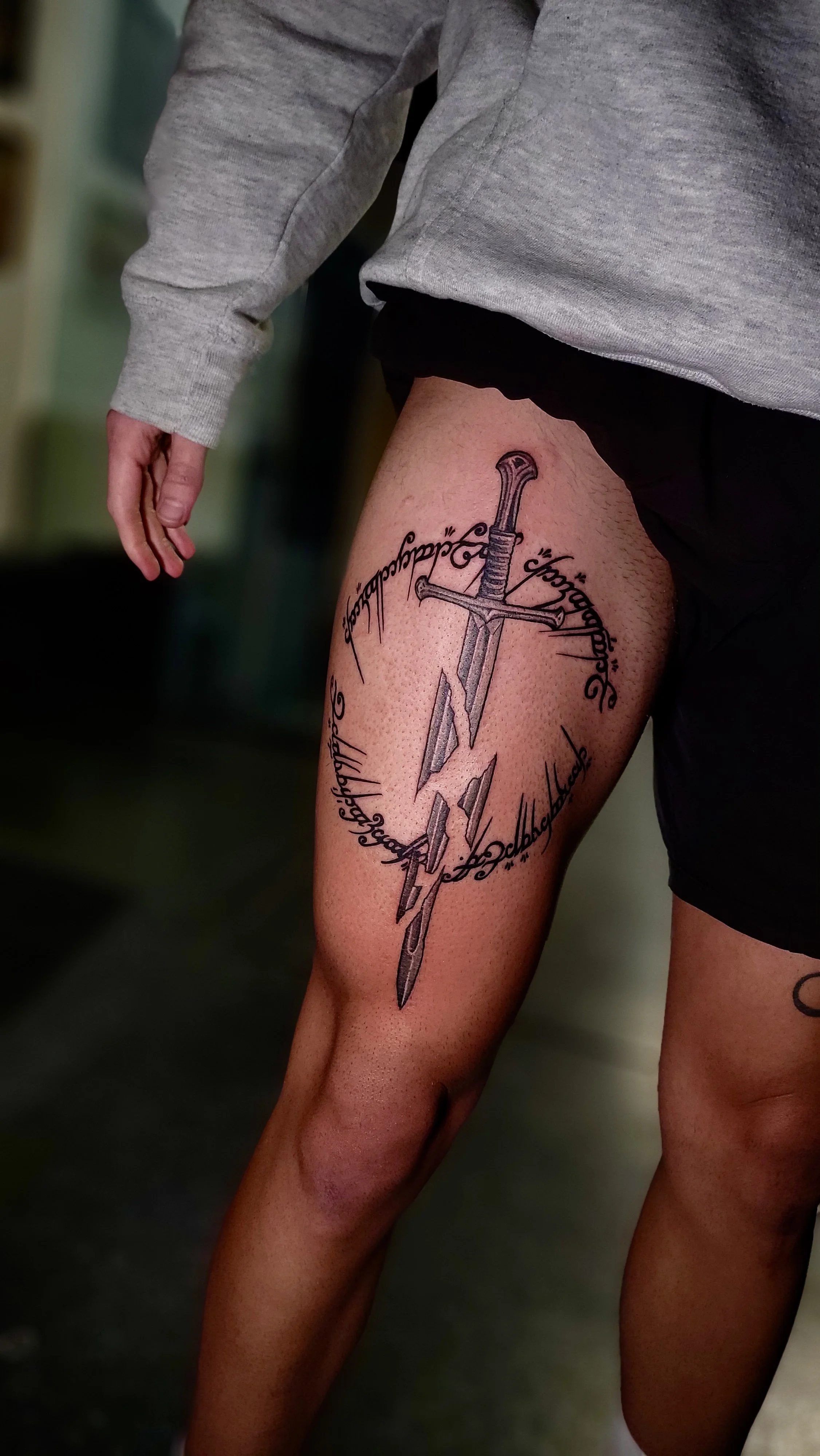 A person's thigh with a detailed black and gray tattoo of a sword, surrounded by cursive writing.