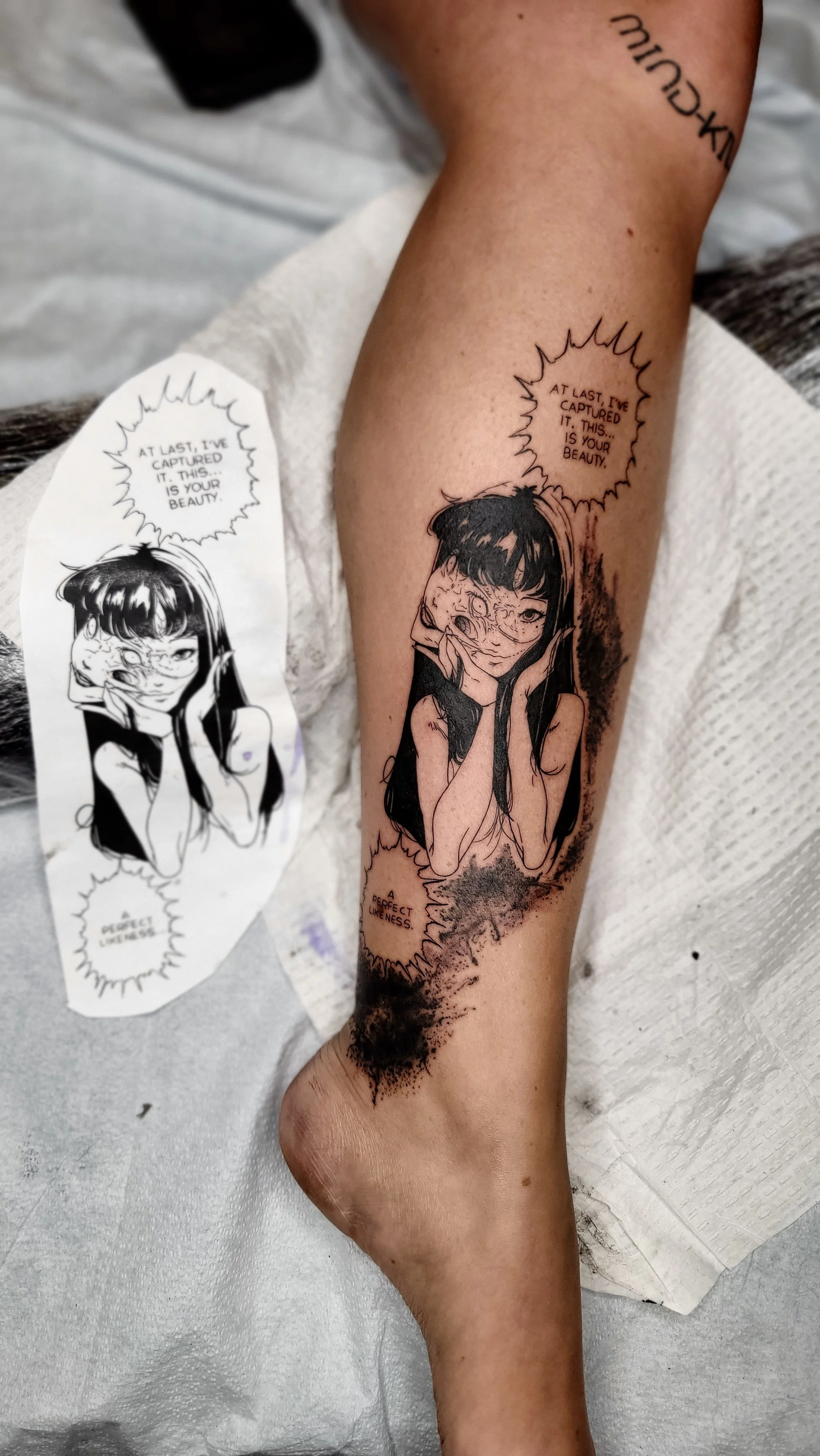 A tattoo of a woman with long dark hair, resting her face in her hands, with a speech bubble that reads 'AT LAST, I'VE CAPTURED IT. THIS... IS YOUR BEAUTY.' The tattoo is on a person's arm, with a drawing of the same woman on a paper nearby.