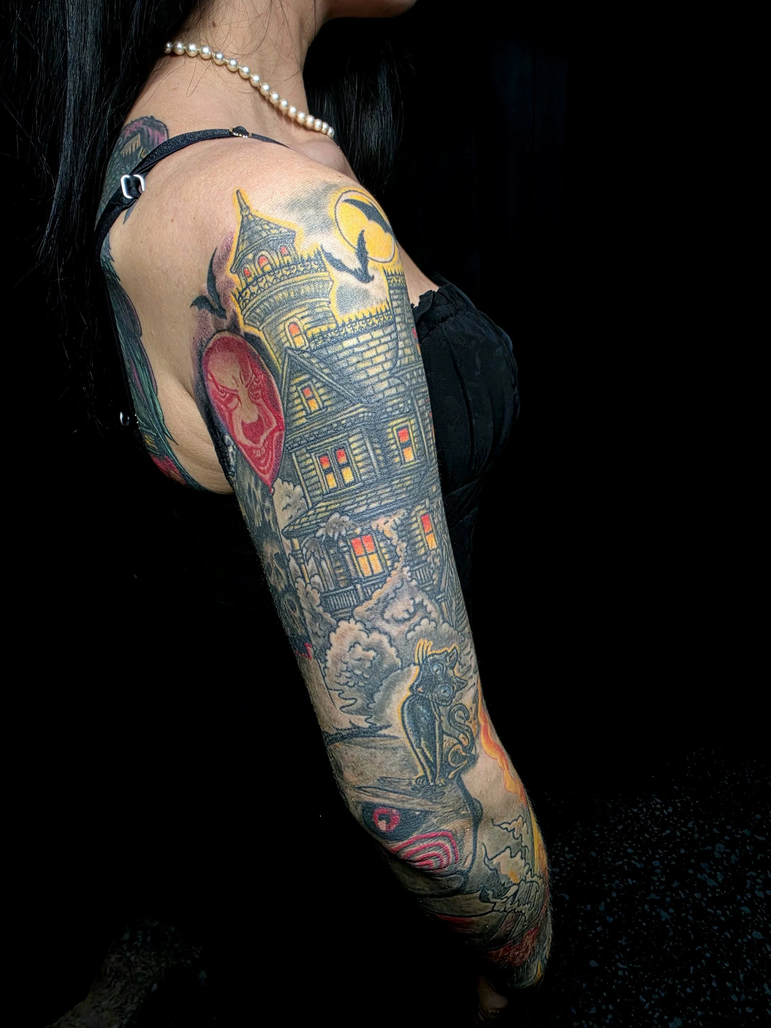 A close-up photo of a person's arm and shoulder, displaying detailed colorful tattoos of a gothic building, a red face mask, a creature with wings, and various other designs, against a black background.