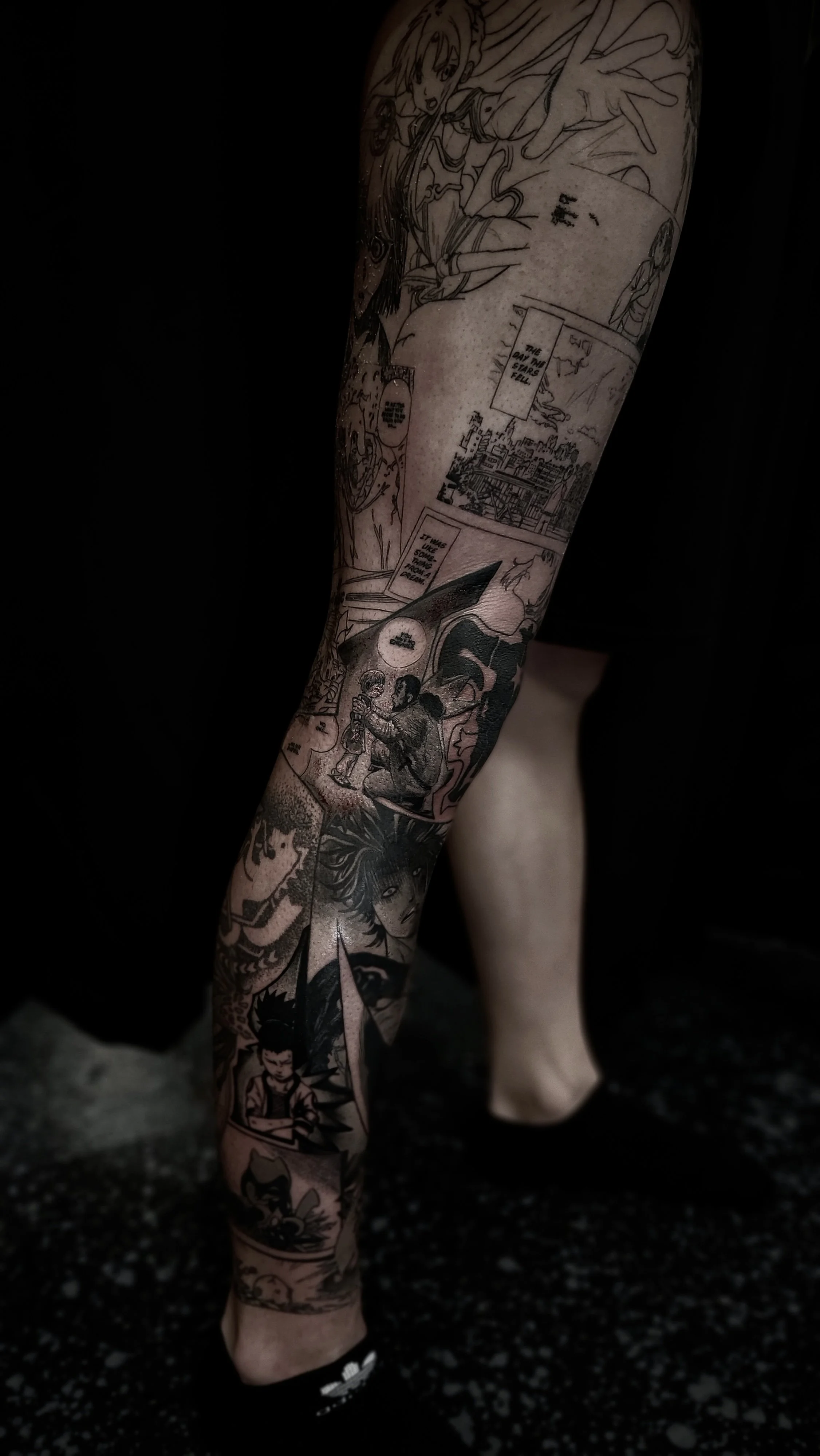 Close-up of a person's arm covered in detailed black and white comic book-style tattoo artwork, featuring various characters, scenes, and speech bubbles.
