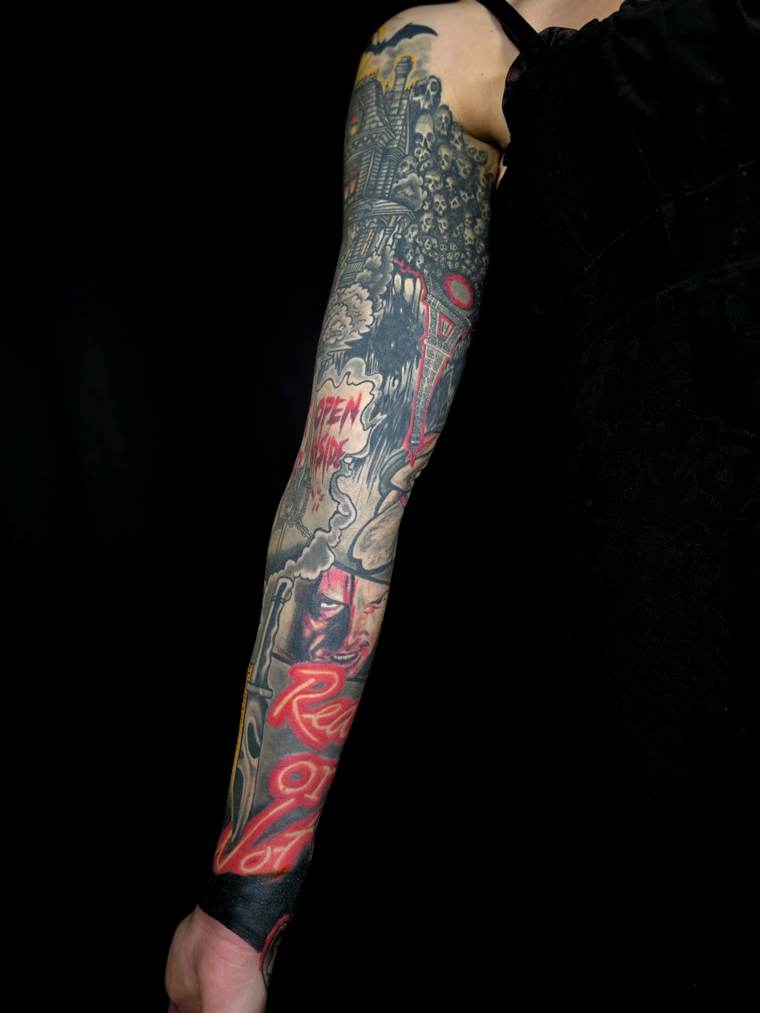 Close-up of a person's arm with extensive colorful tattoos, including skulls, a house, clouds, smoke, and text reading 'OPEN WIDE' and 'RESIST' against a black background.