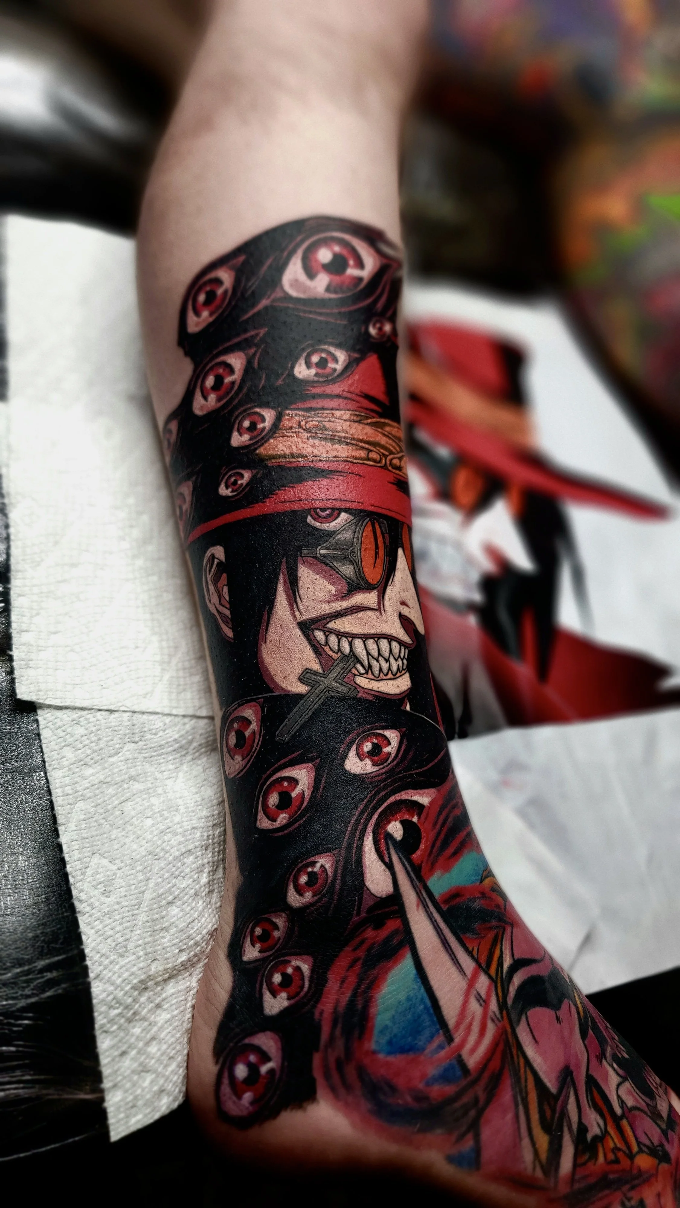 Tattoo of a smiling skeleton with orange sunglasses and a cross necklace, surrounded by red eye motifs, on someone's arm.