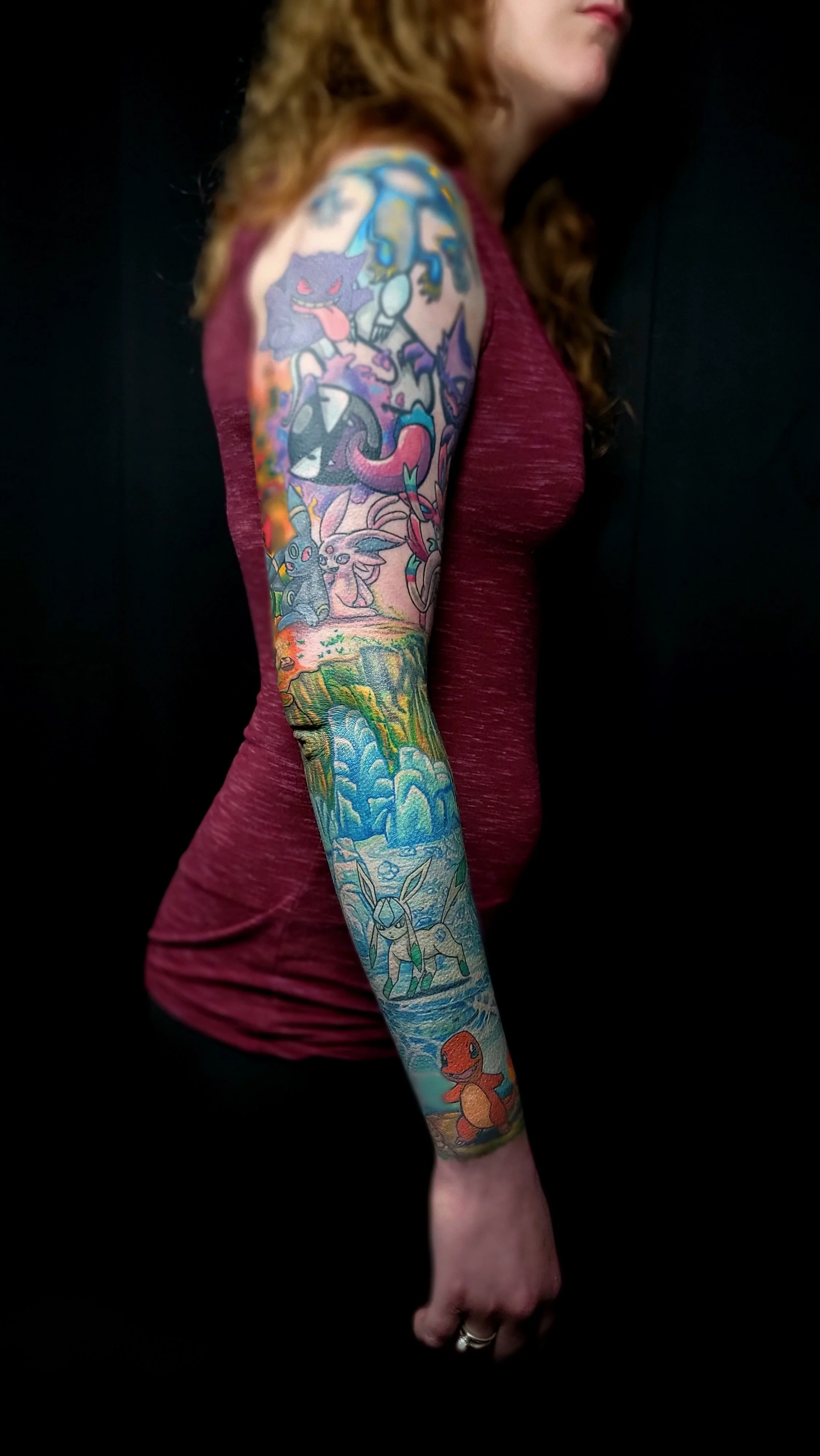 A woman with long curly hair wearing a maroon sleeveless shirt showing a colorful full sleeve tattoo with various cartoon characters and scenes, standing against a black background.