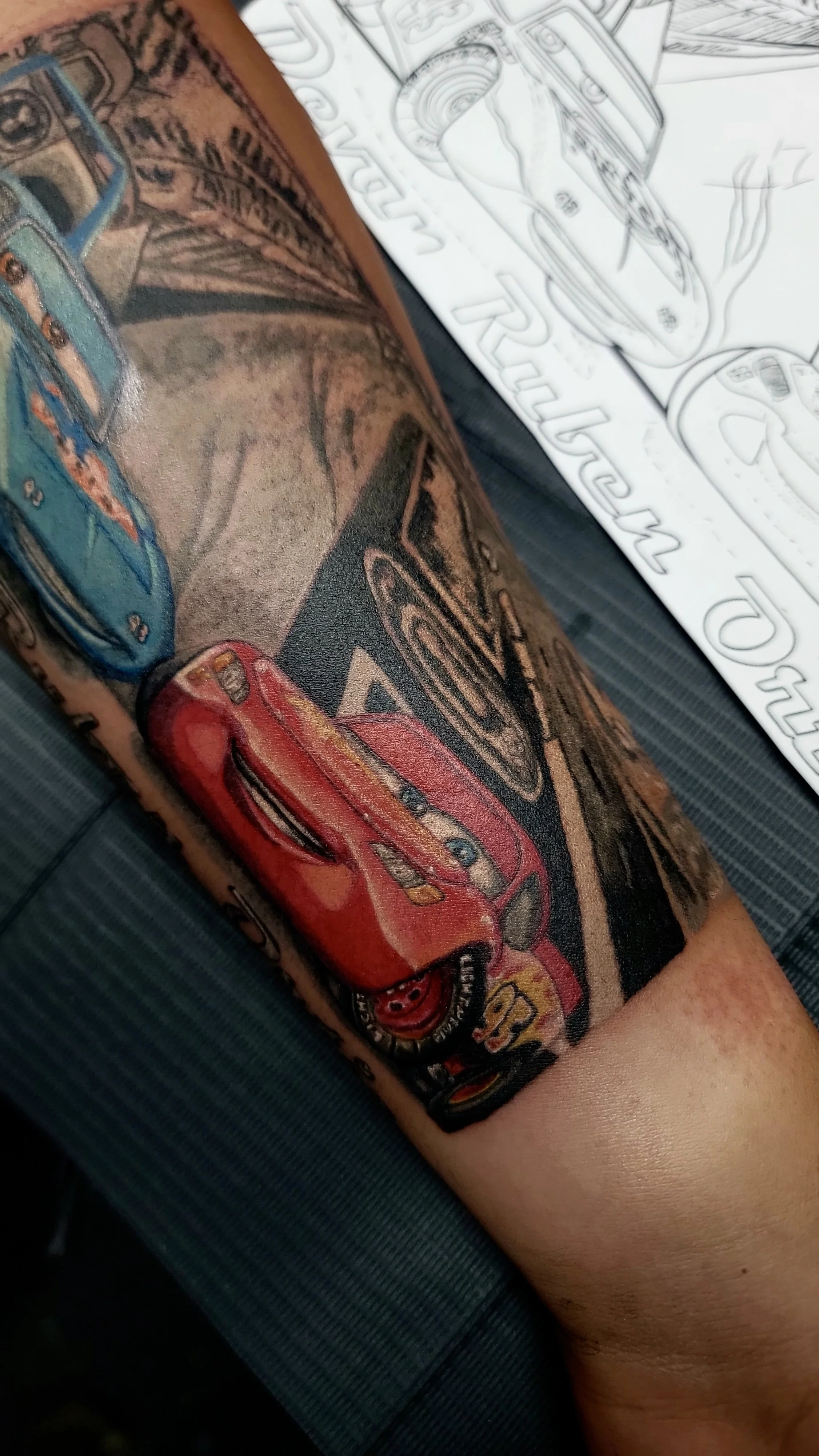 Colorful tattoo of race cars on a person's arm with a sheet of car coloring pages in the background.