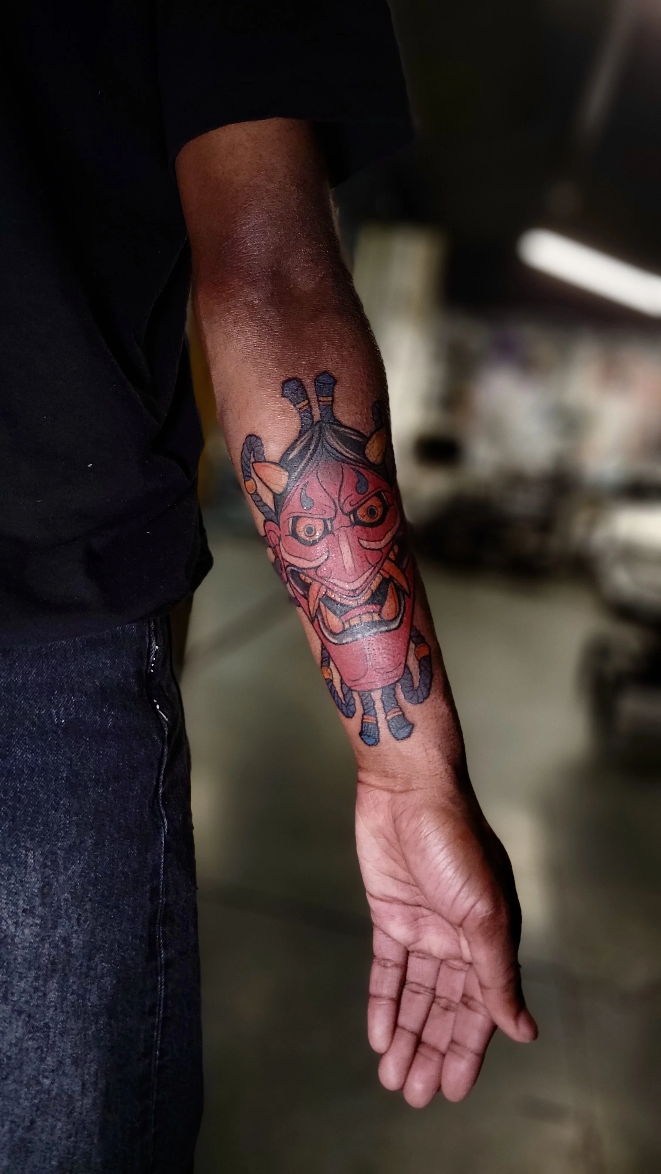 A person's arm with a colorful traditional-style tattoo of a face and a snake, and the person's hand extended downward.