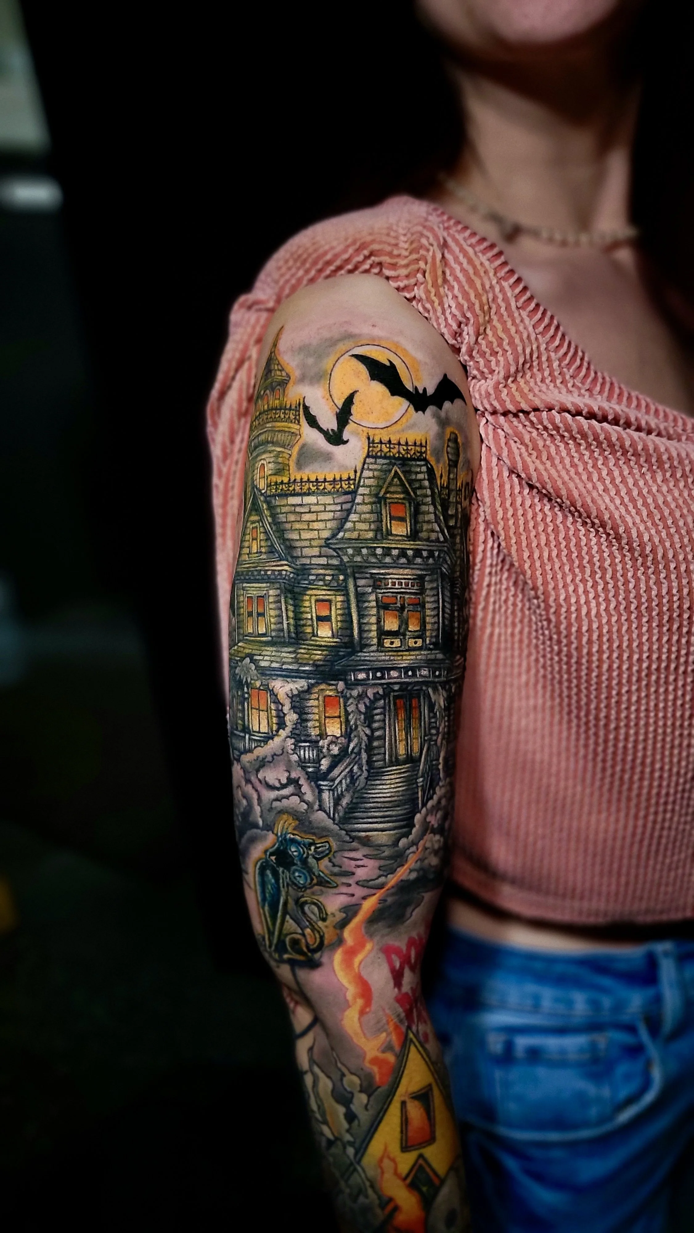 Tattoo of a haunted house with bats flying in front of a large moon, a witch on a broomstick, and a horse with a flaming tail on a person's upper arm.