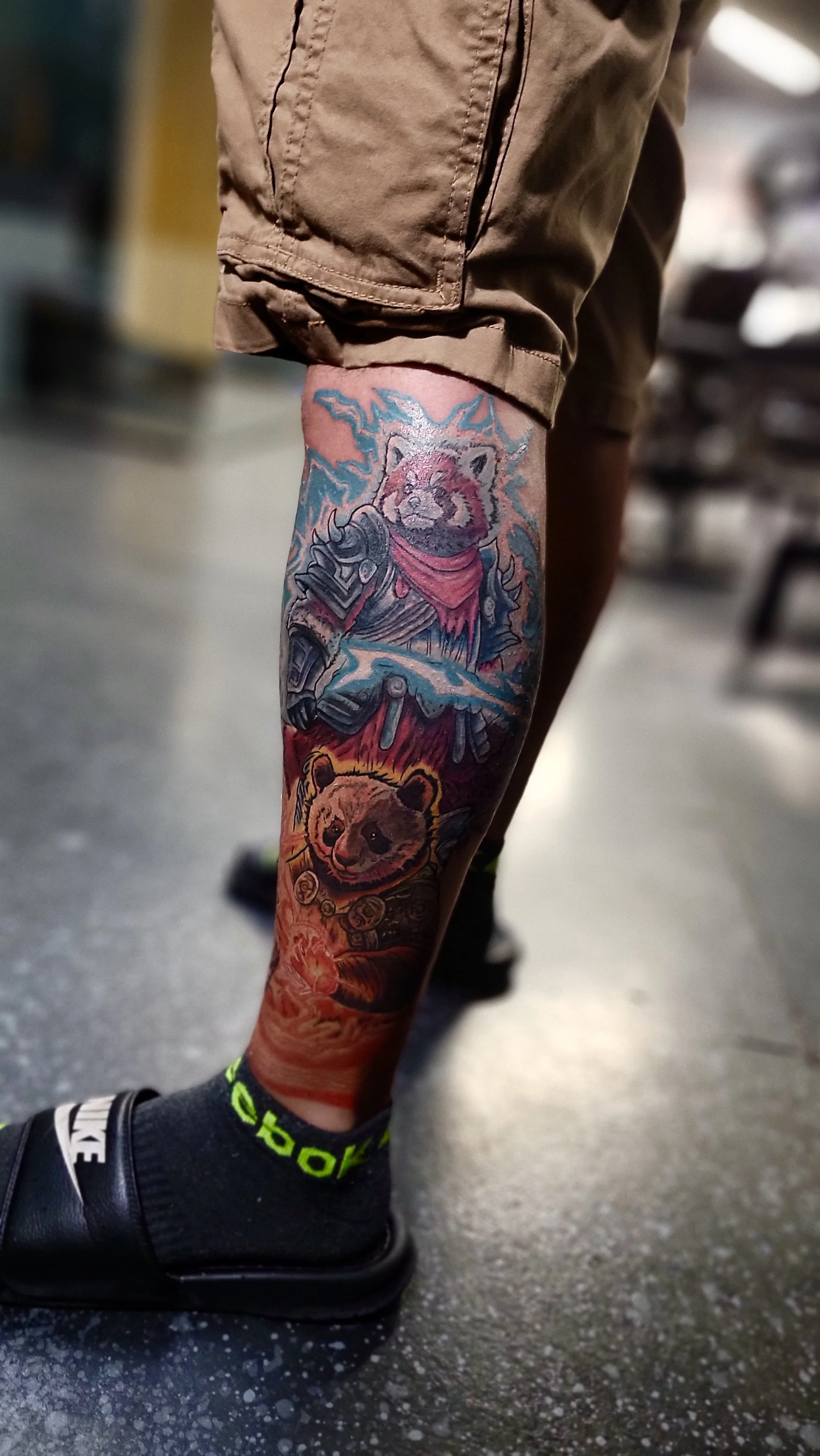 Tattooed leg with a bear cub and lightning bolt design, wearing a brown short sleeve shirt, black Nike sneakers, and gray socks with bright green accents, on a speckled gray floor.