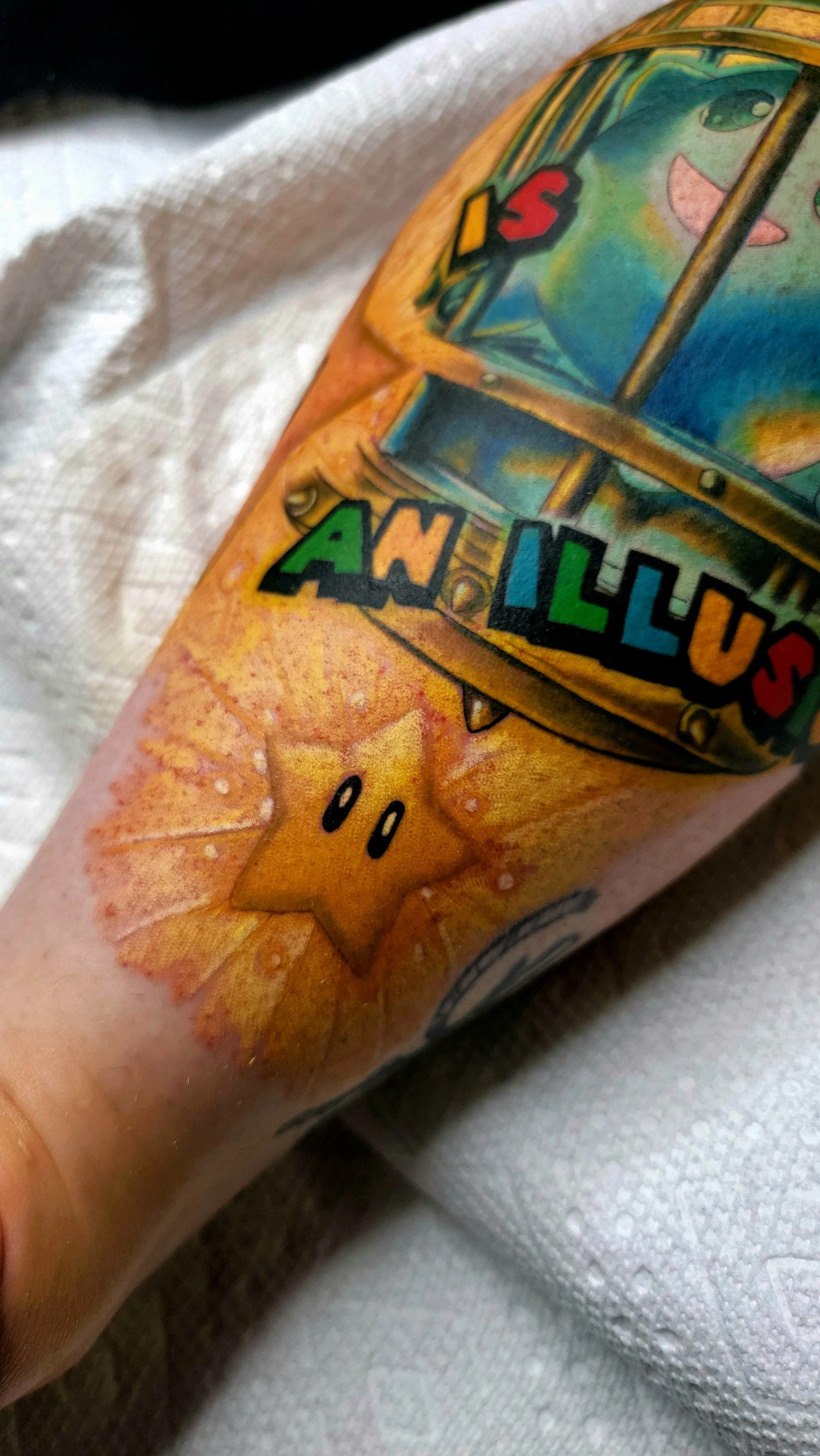 Close-up of a tattoo featuring a yellow star with a smiling face on a person's arm, with colorful cartoon-style artwork including a pinball machine with the words 'IS' and 'A WORLD' on it.