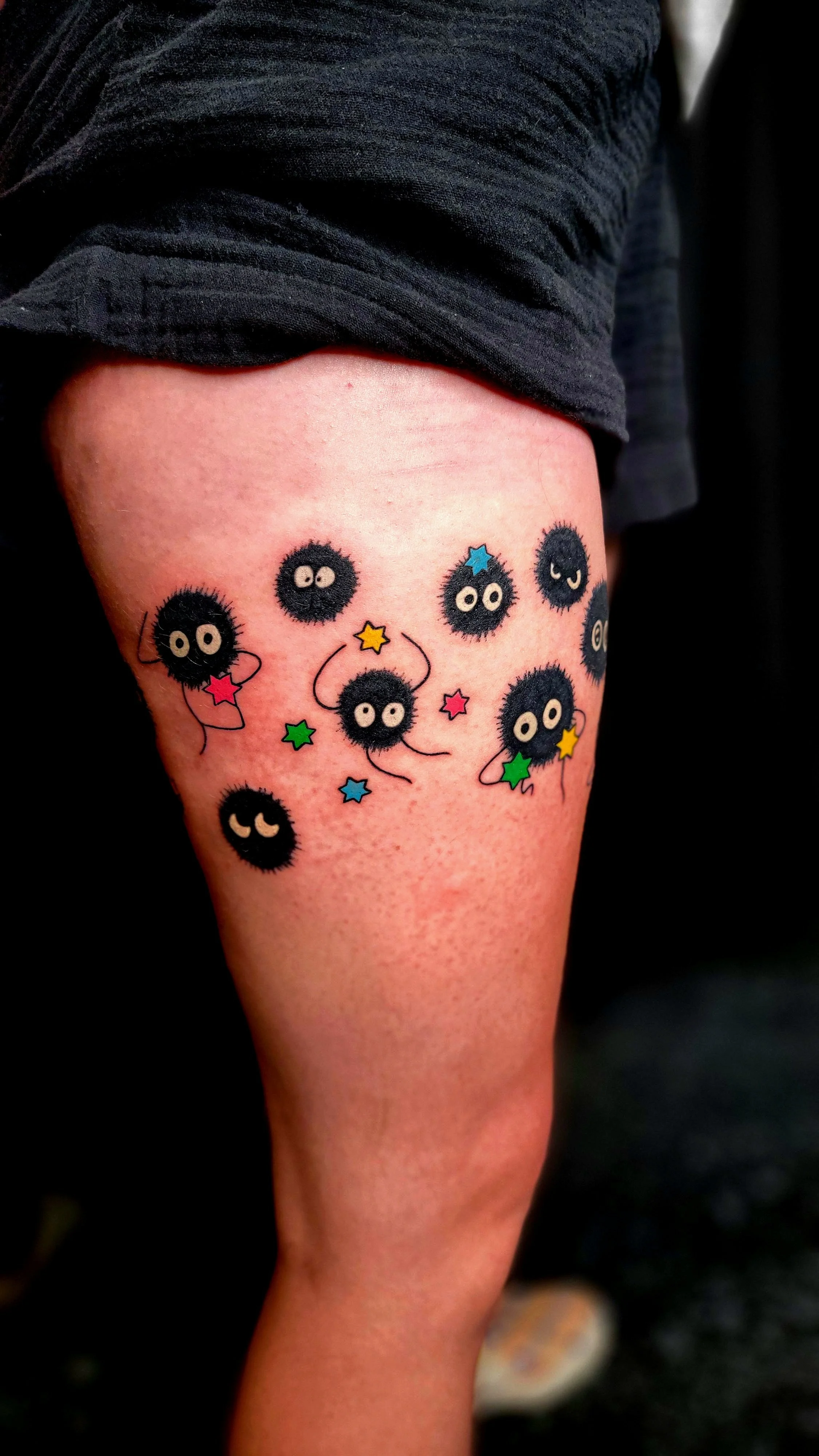 A tattoo on a person's thigh featuring cartoonish black fuzzy balls with eyes, some with facial expressions, and colorful stars and squiggly lines.