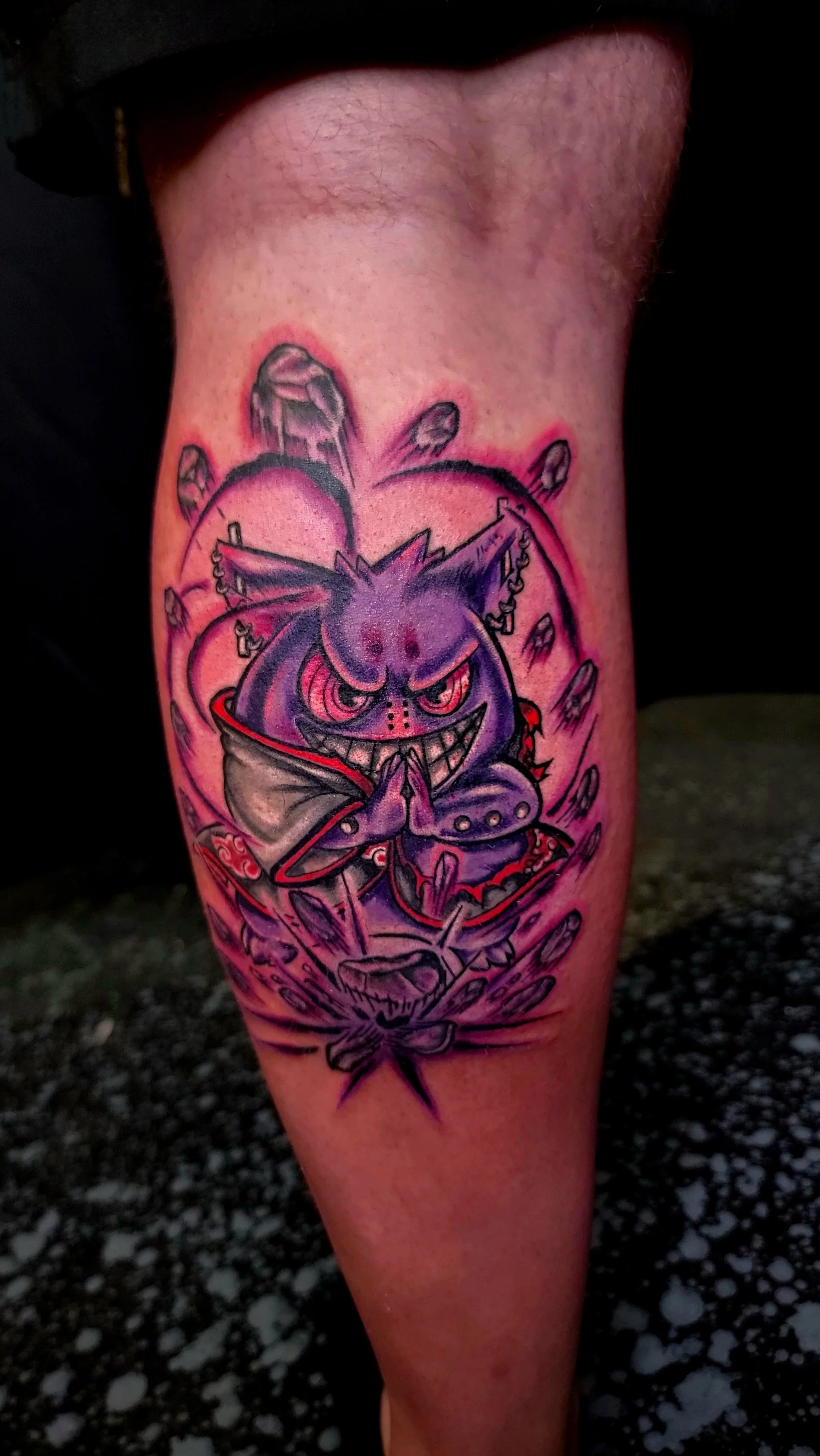 Colorful tattoo of a mischievous purple cartoonish creature with pointy ears and a wide smile, seated with hands clasped, surrounded by a chaotic background of feathers, objects, and abstract designs.