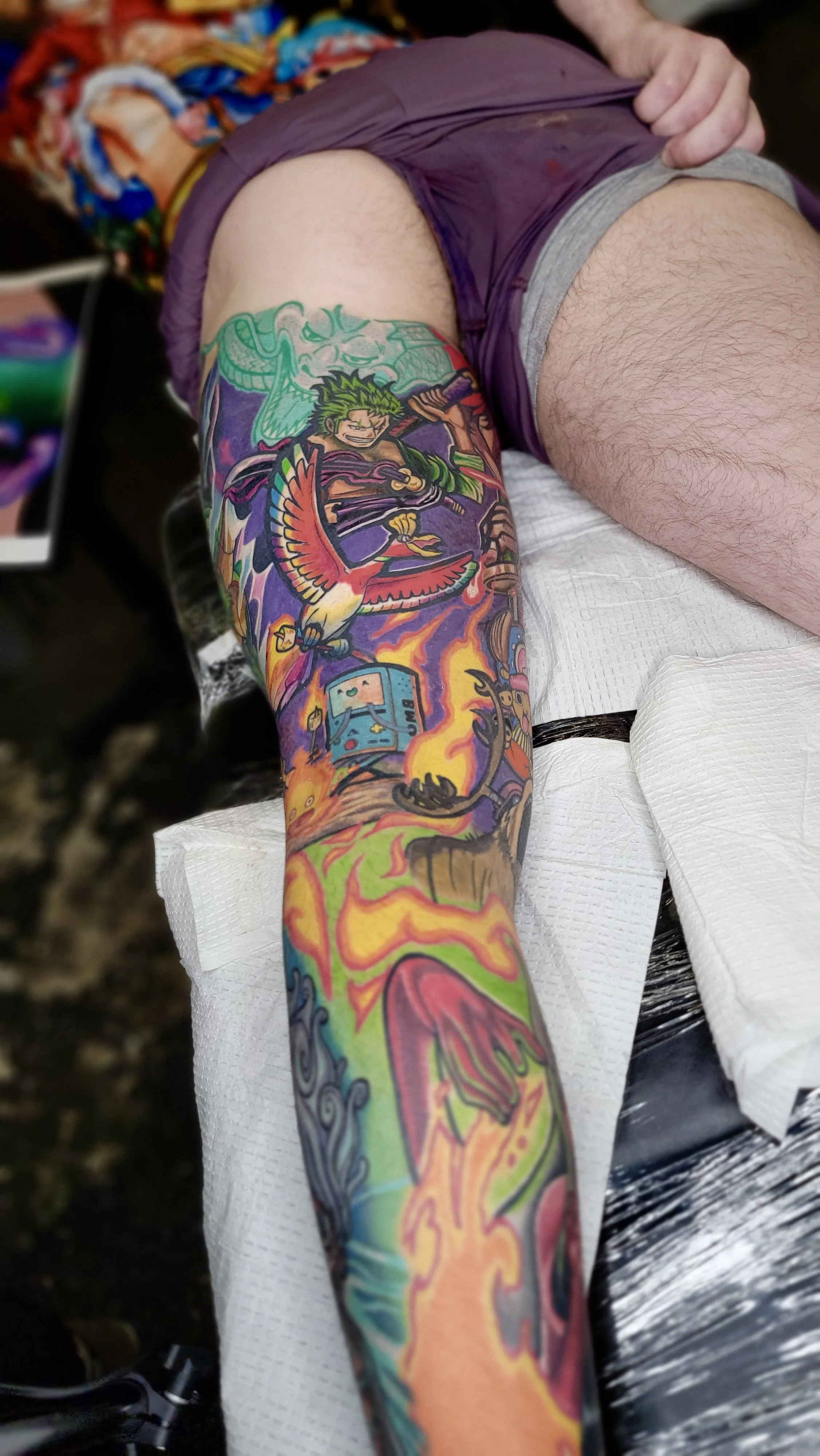 Colorful tattoo sleeve featuring a character wielding a sword, a bird, a television, and various abstract designs covering a person's arm.