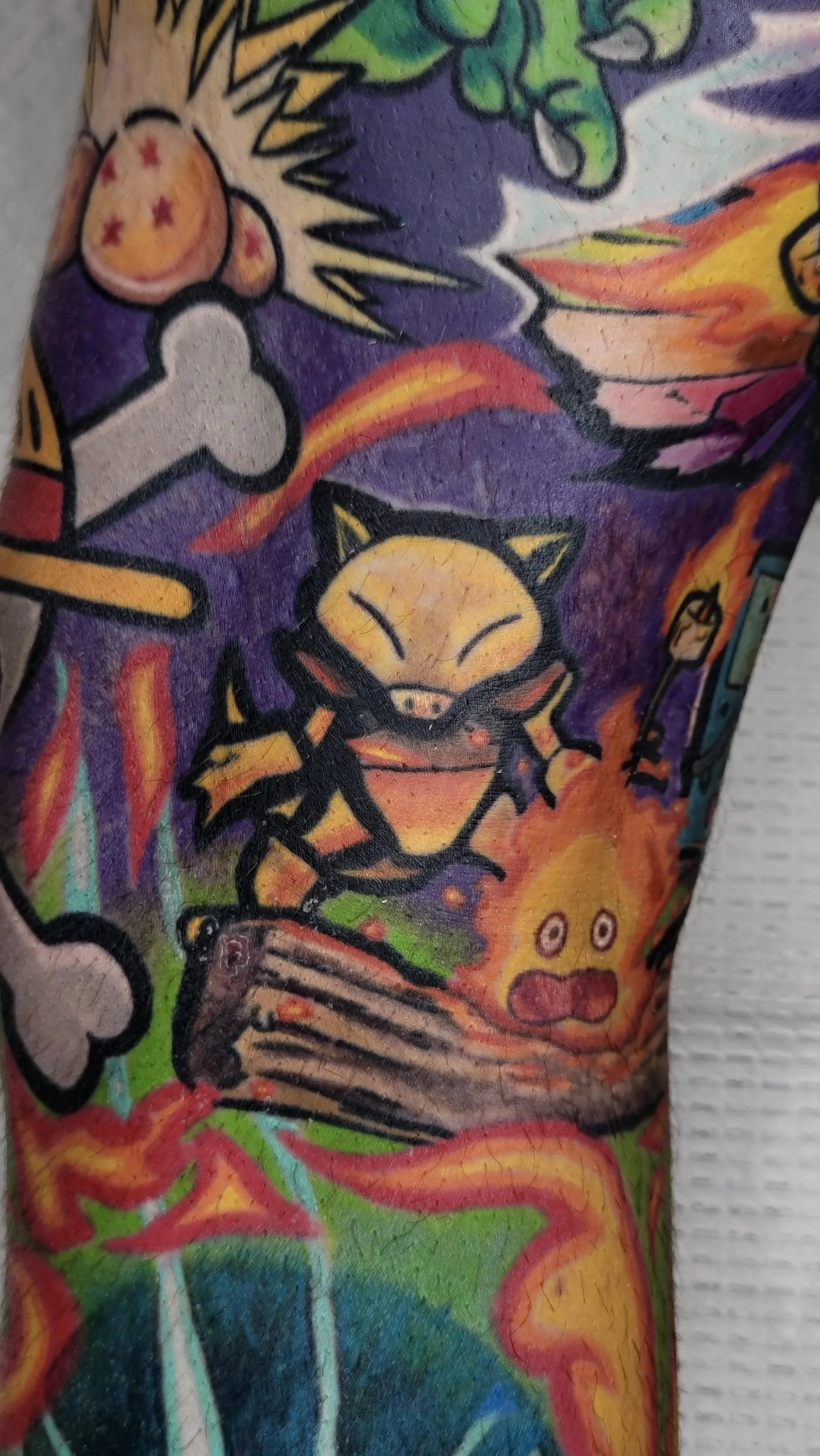 Colorful tattoo featuring various characters, including a smiling cat, skull, flaming objects, and abstract shapes.