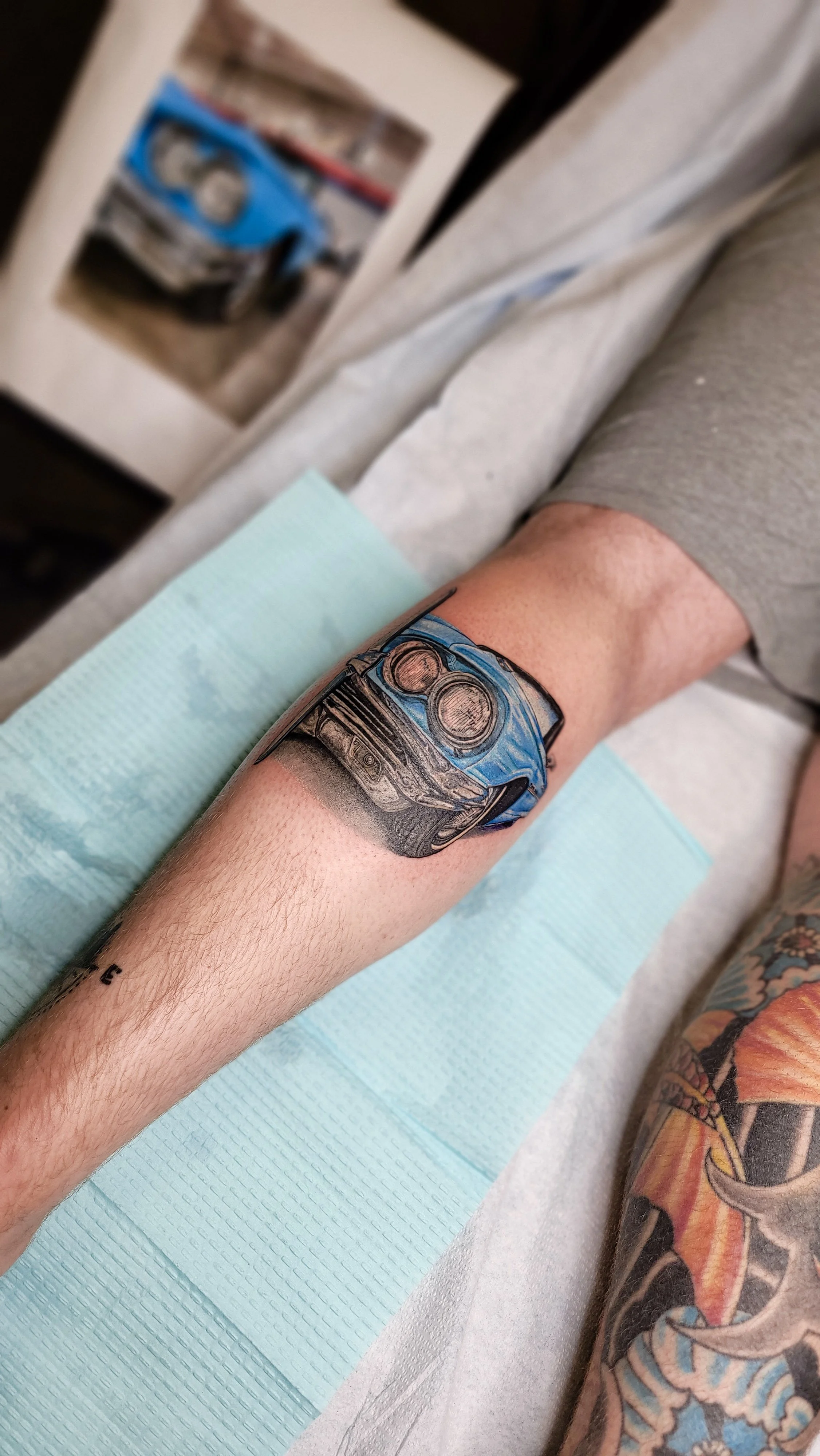 Tattoo of a blue sports car on person's forearm, with part of a colorful sleeve tattoo visible on lower arm.
