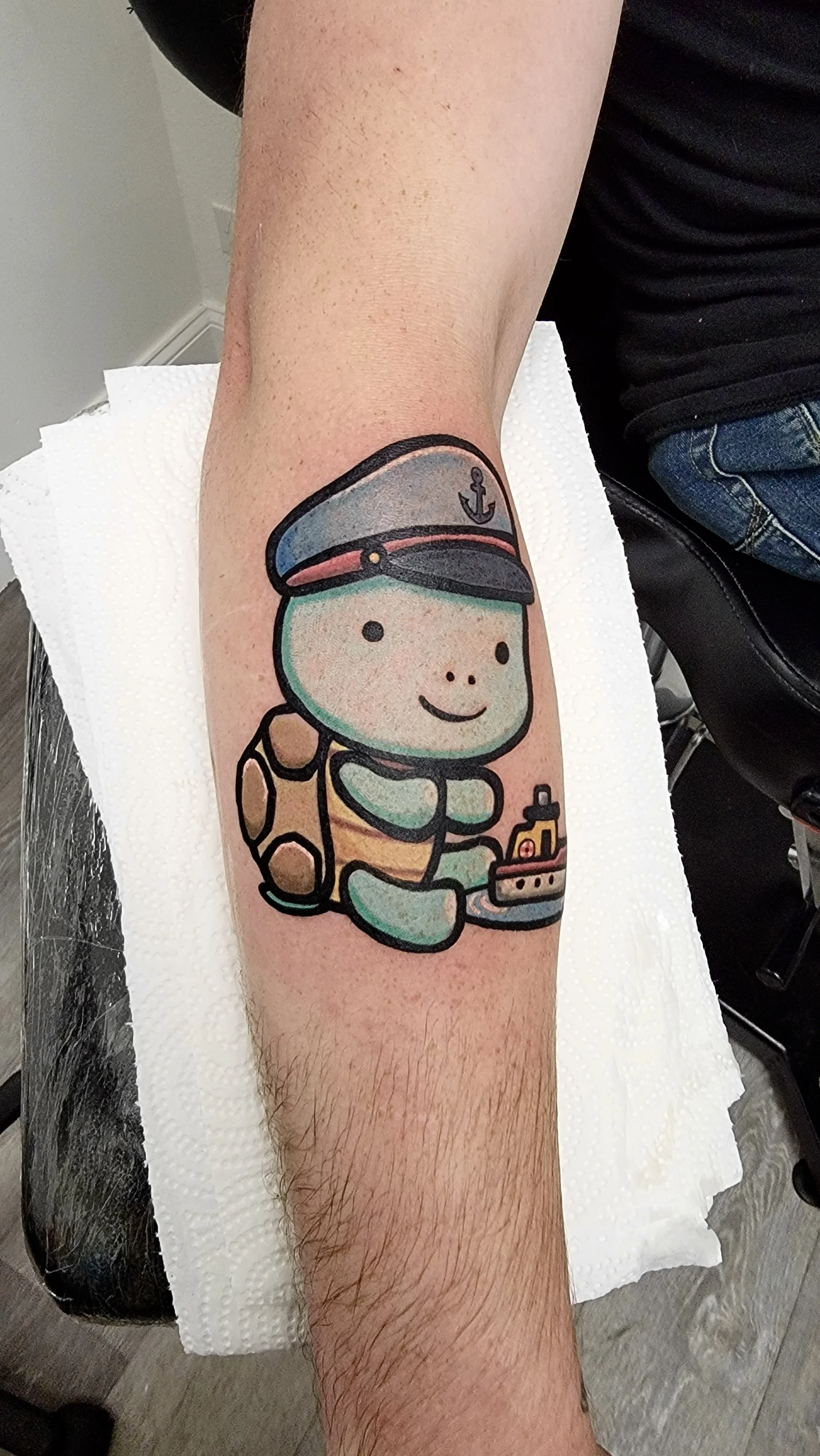 Colorful tattoo of a cute turtle wearing a captain's hat, sitting with its front paws together, holding a small boat, on a person's forearm.