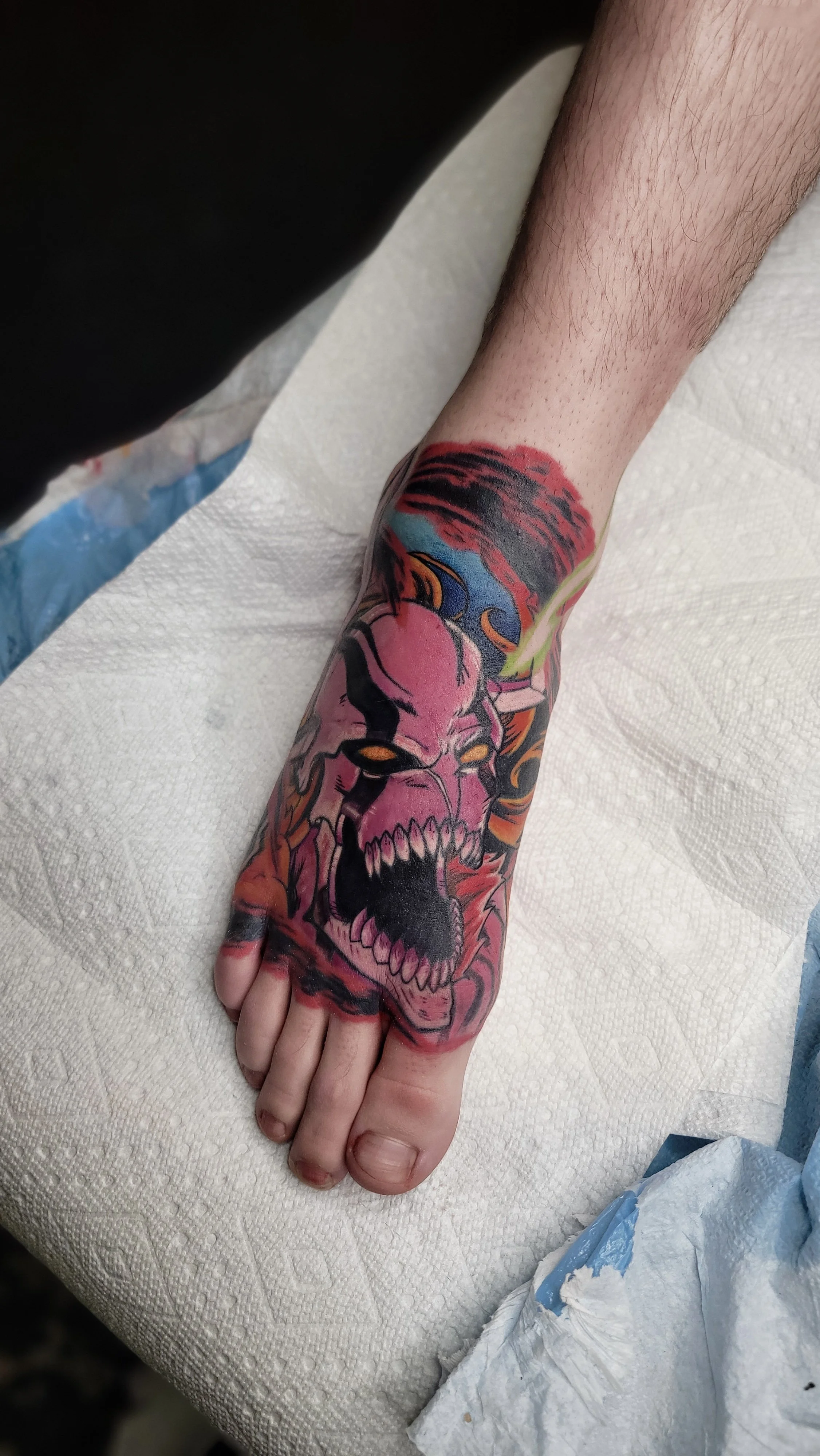 A tattoo of a menacing pink skull with sharp teeth, yellow eyes, and flames, covering the person's forearm and hand.