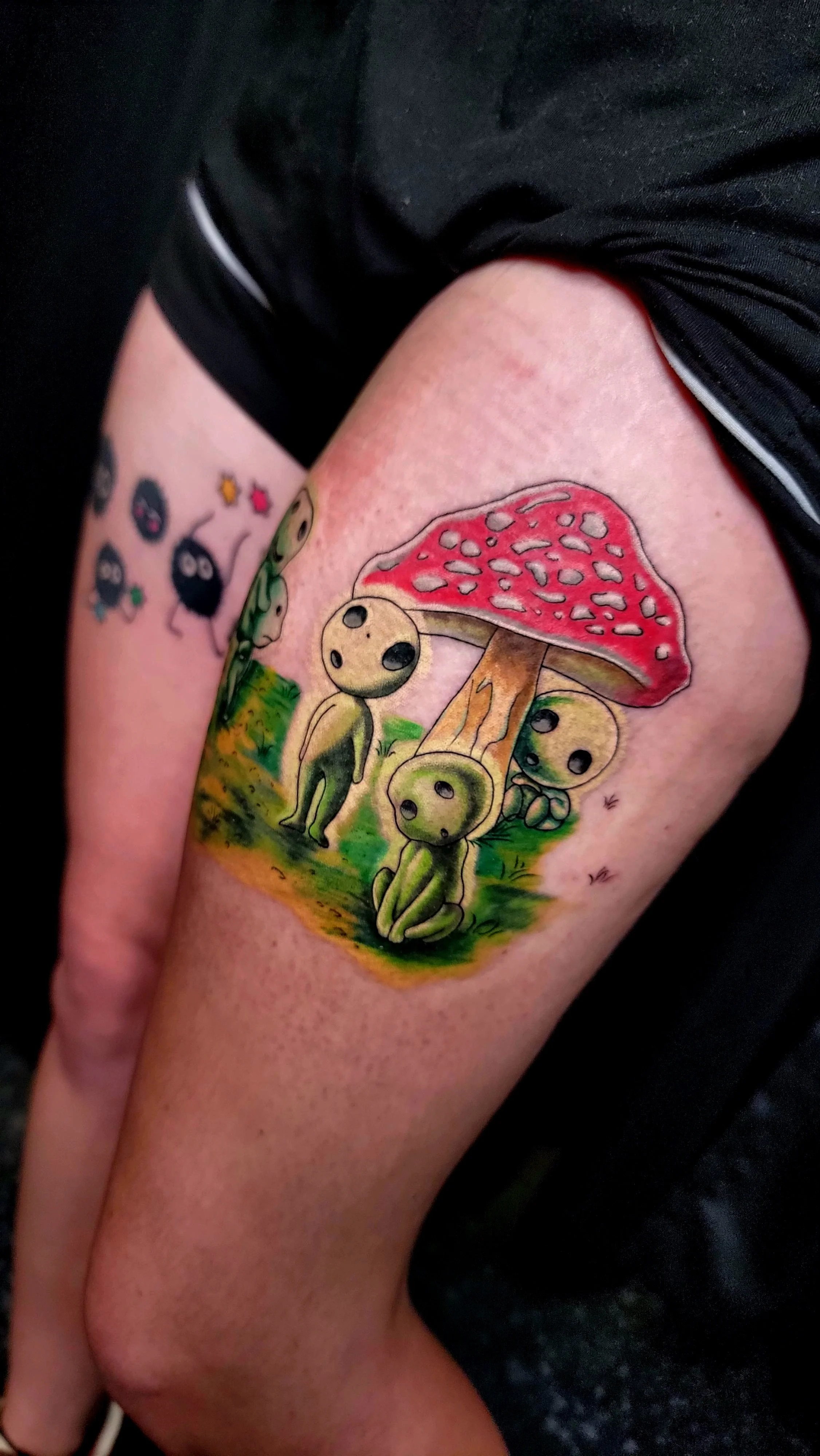 Colorful tattoo of cute green aliens with big black eyes sitting and standing under a red mushroom with white spots on a person's upper arm.