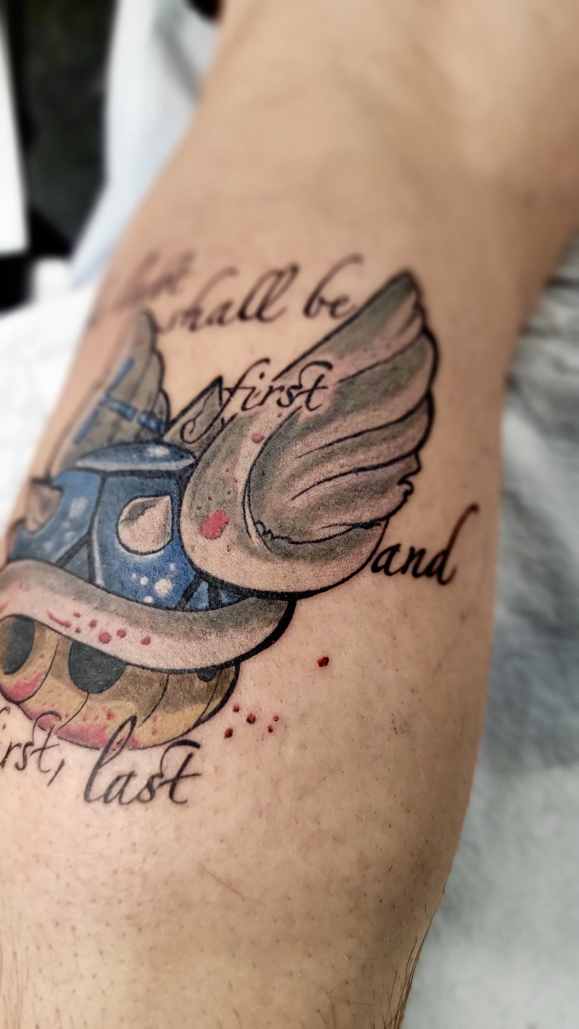 Tattoo on skin that reads 'smallest and last' with a colorful angel wings and a shield design.