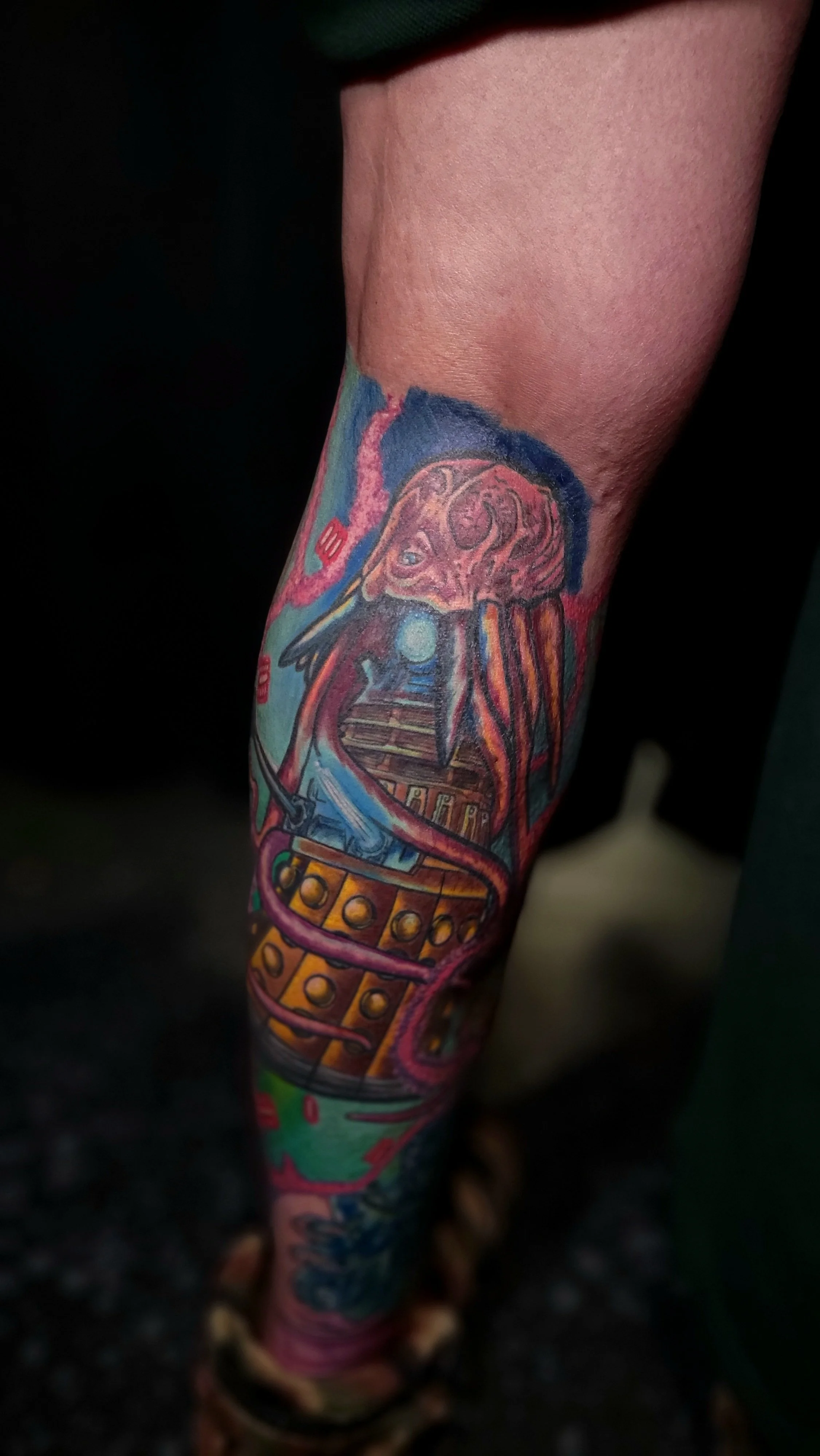 Colorful tattoo of a creature with tentacles, a large eye, and a fire logo on its head on a person's arm.