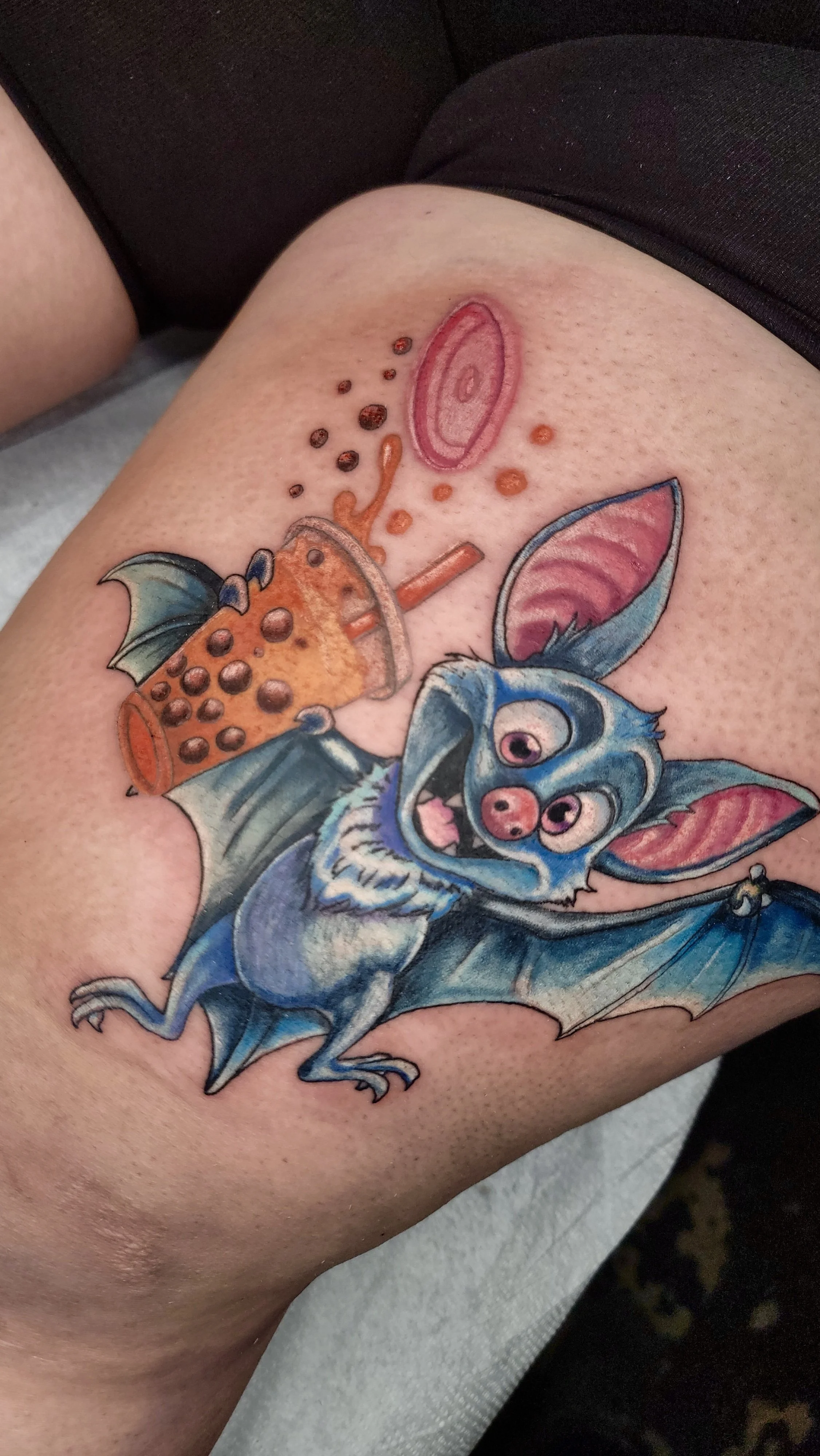 Colorful tattoo of a cartoon bat with large eyes, pink face, and wings, holding a small purple object, on person’s upper thigh.