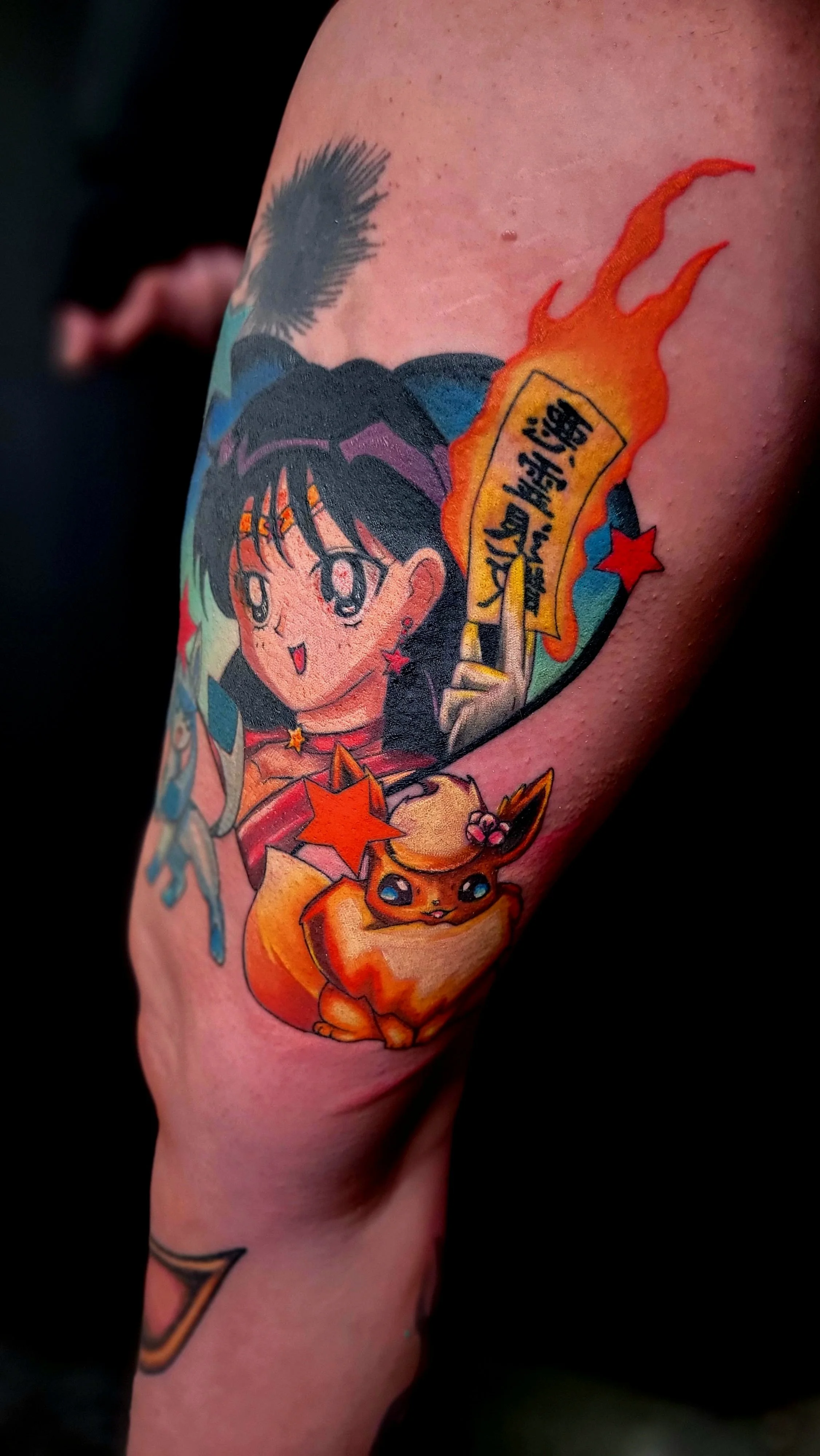 Colorful tattoo on person's arm featuring an anime-style girl with black hair, a small yellow creature resembling Eevee from Pokémon, and a fiery speech bubble with Japanese text.