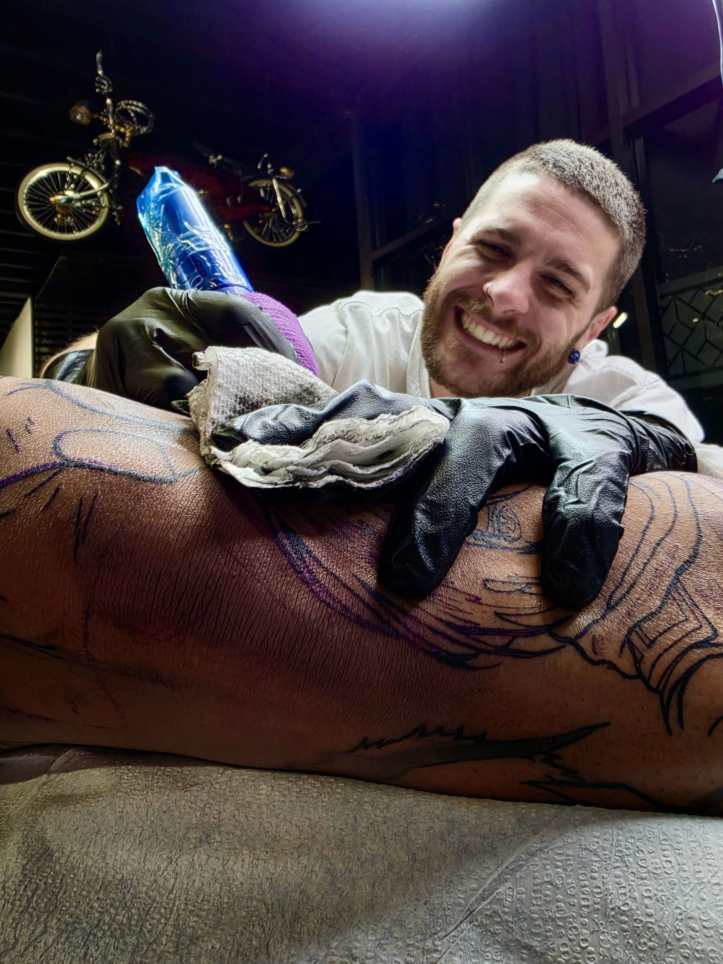 A man with a beard and short hair is smiling while tattooing a person's leg. The tattoo includes colorful designs, and the tattoo artist is wearing gloves. In the background, a decorative bicycle and some dark walls are visible.