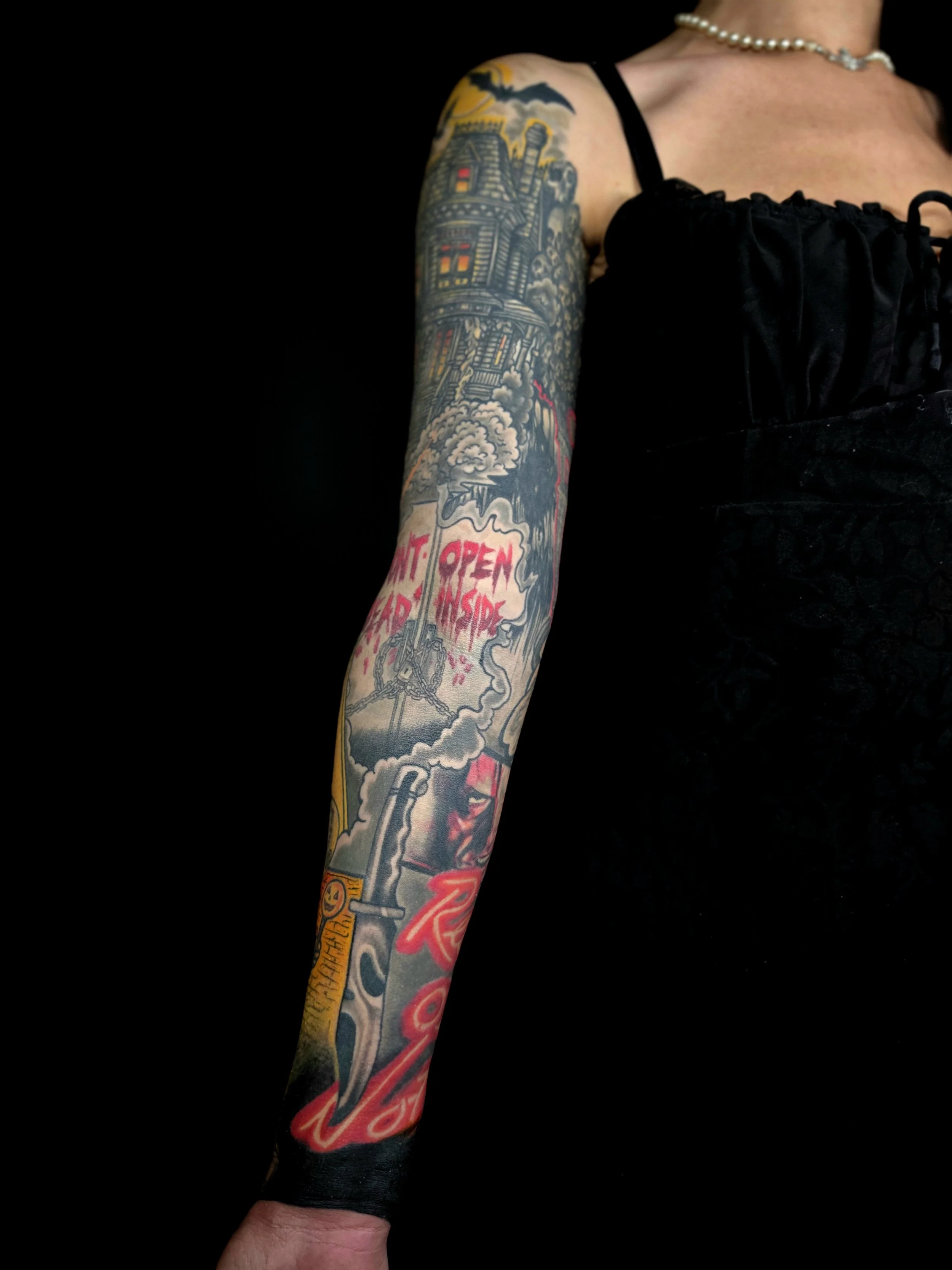 Person with colorful tattoos on their arm, wearing a black dress with spaghetti straps, a pearl necklace, and holding a black glove.