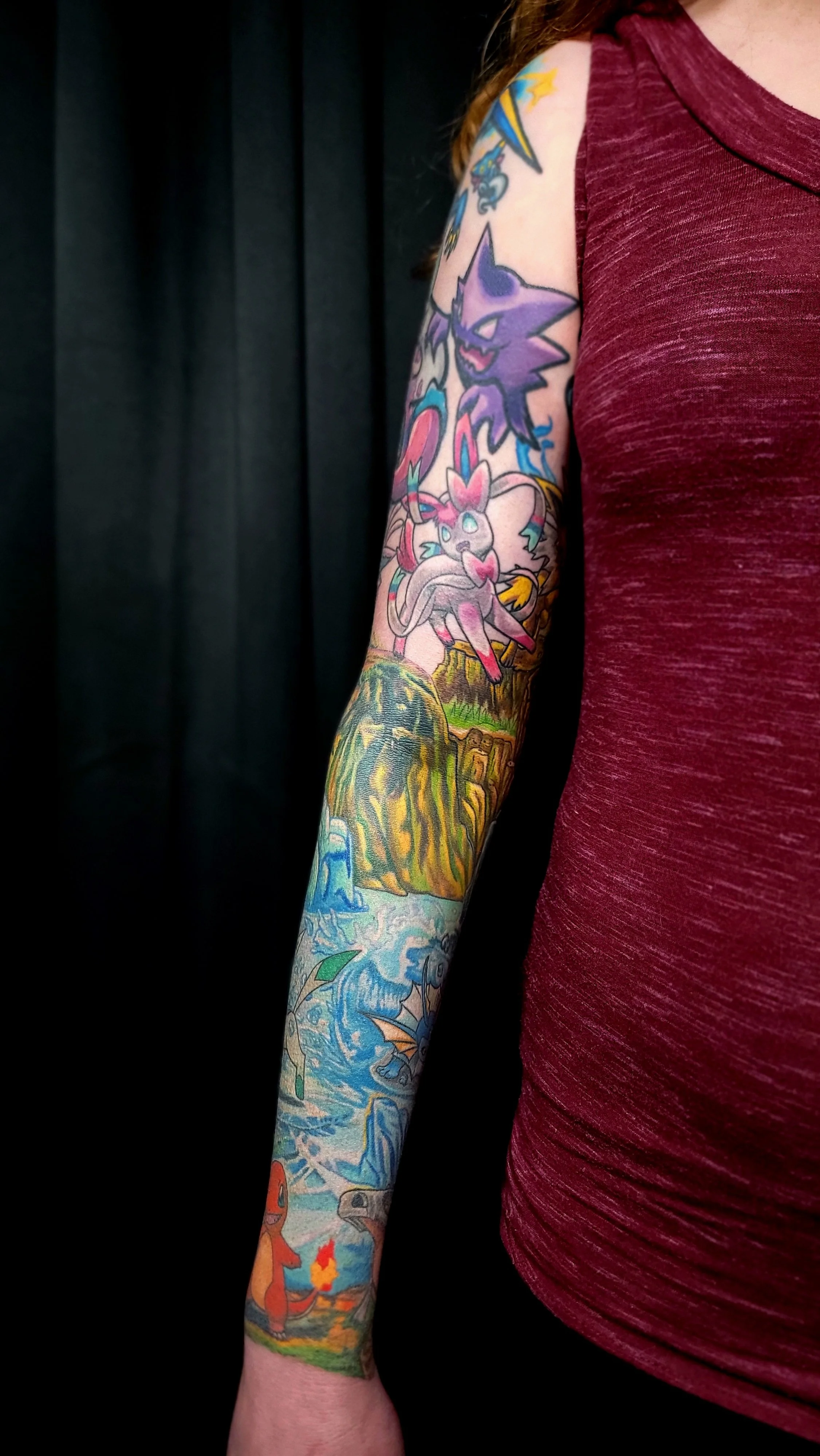 A person's arm with colorful tattoo sleeve featuring cartoon characters, animals, and nature scenes, wearing a red shirt.