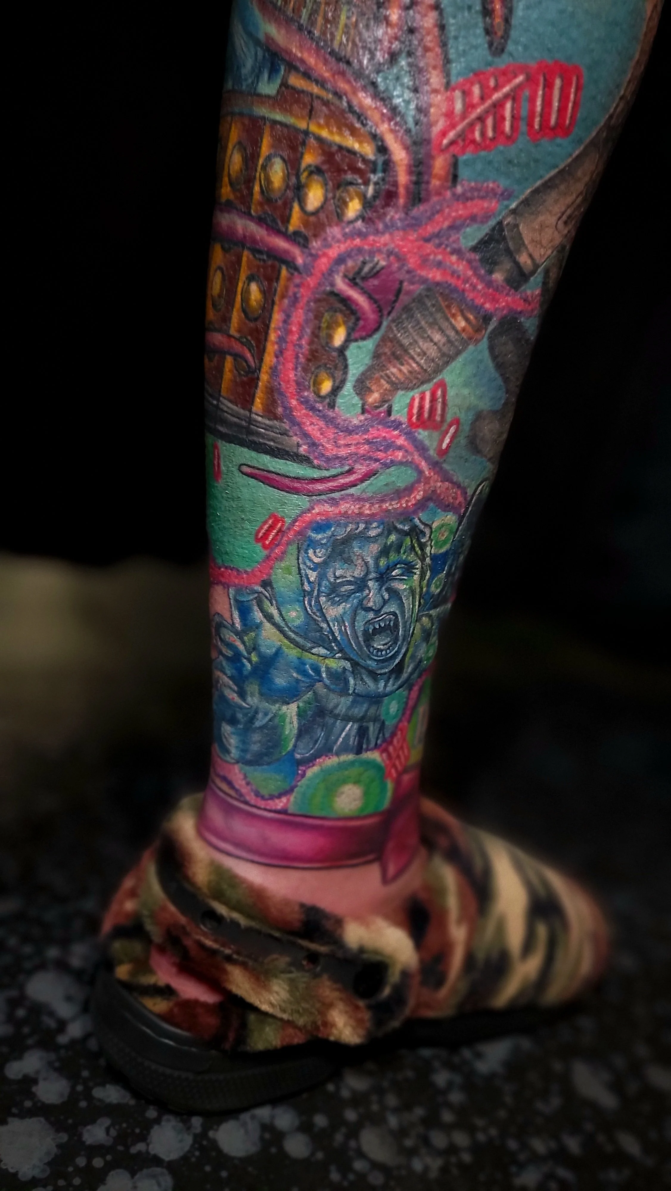 Close-up of a person's leg with colorful tattoos, including a screaming face, a pink octopus, a building, and other designs, wearing a fuzzy multicolored boot.