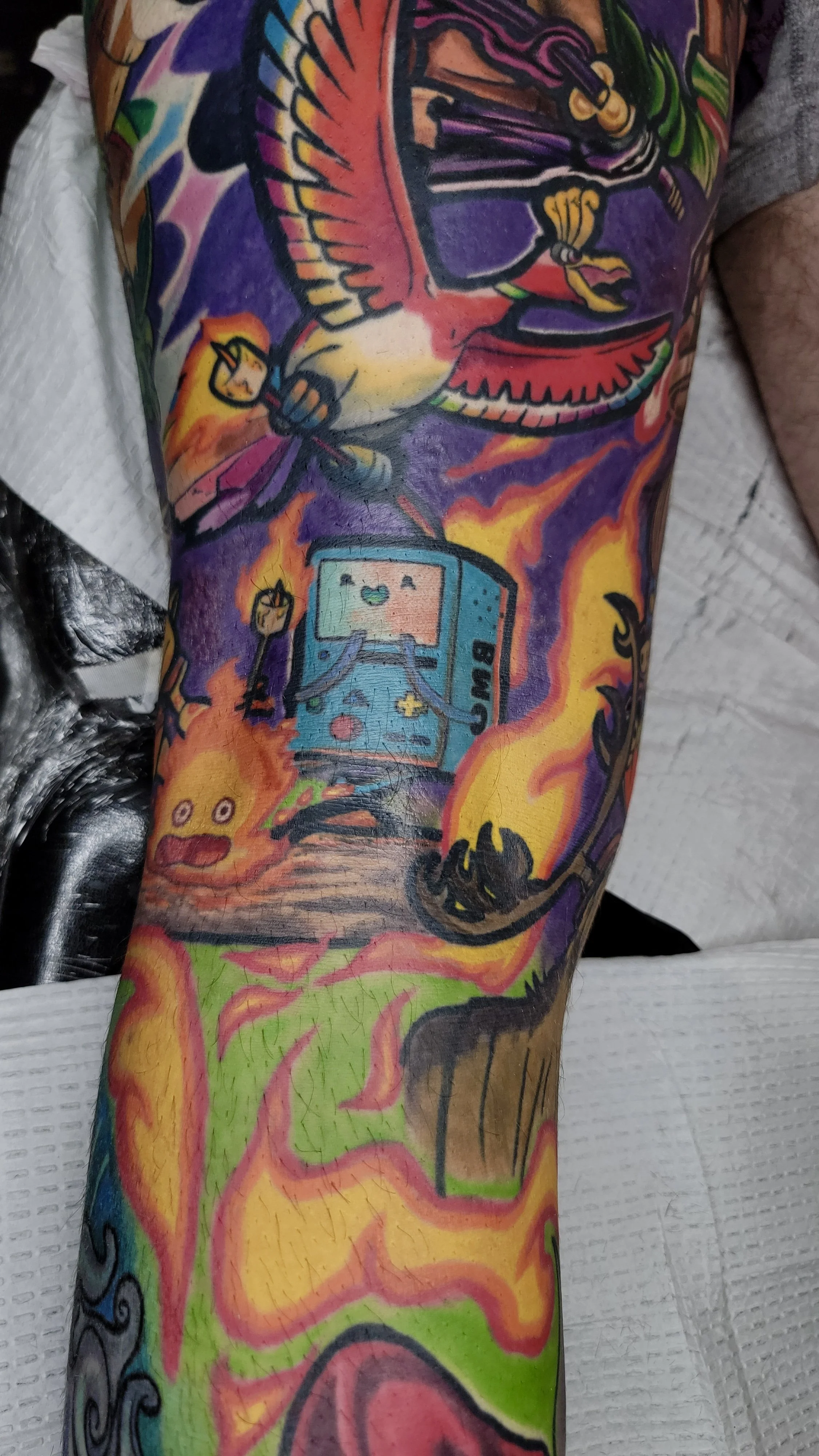 Colorful tattoo on a person's arm featuring a bird, a video game controller, a smiling emoji, and other vibrant, abstract designs.