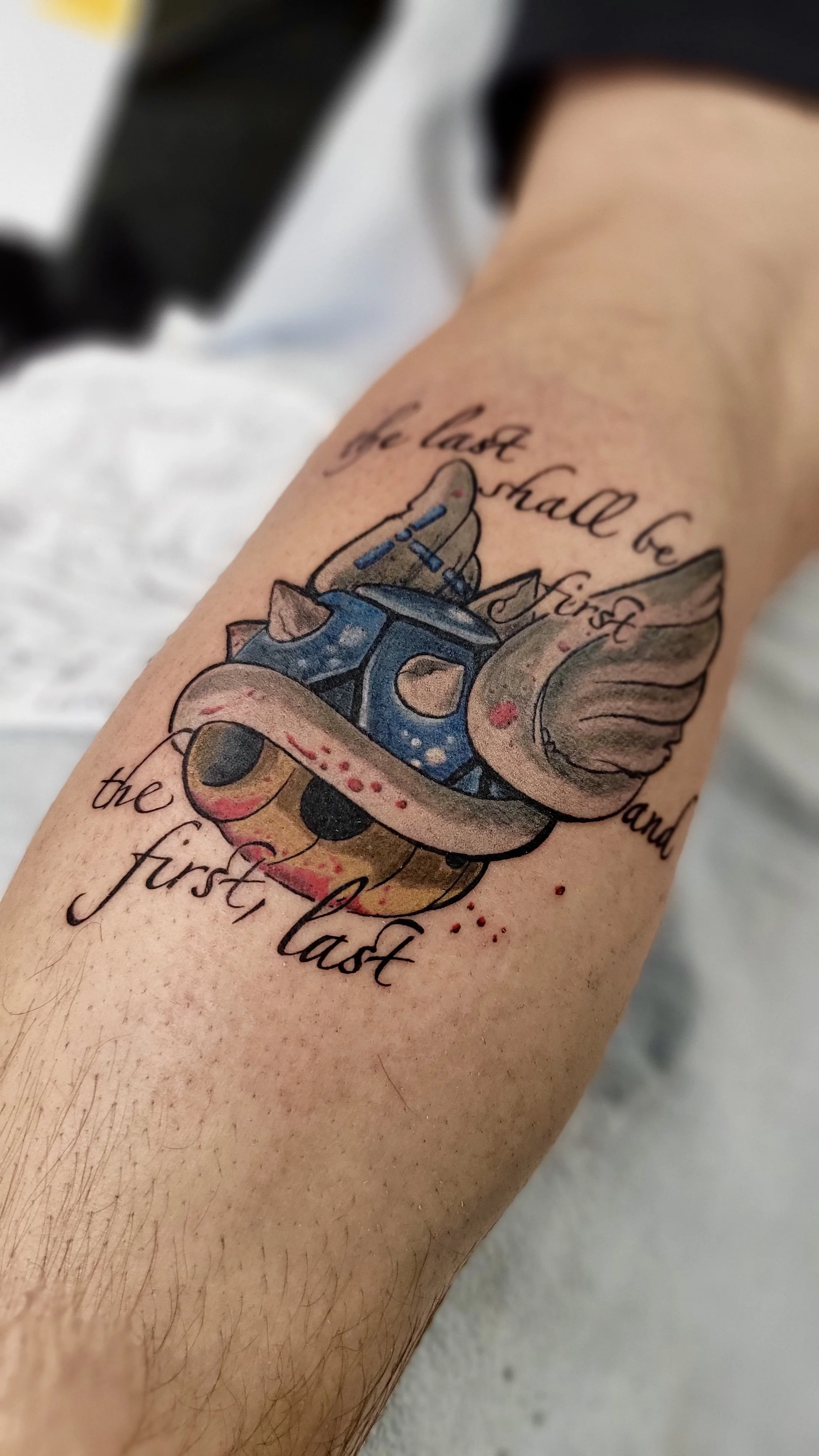 Tattoo on forearm of a cupcake with a skull face, a spoon, and wings, with the quote "the last shall be first and the first last" written around it.