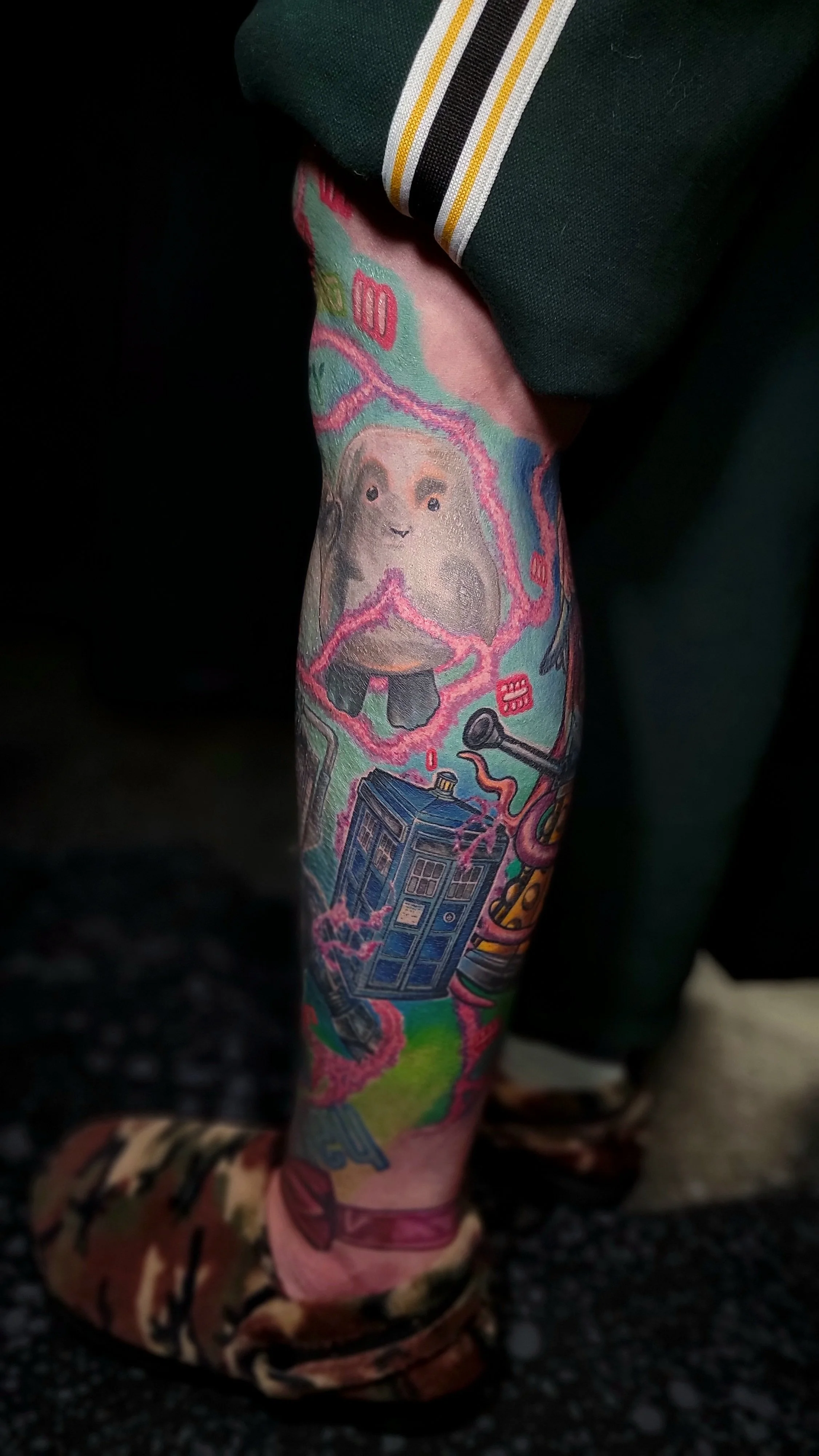 Close-up of a person's arm with colorful tattoos featuring Doctor Who elements, including the TARDIS and a dalek, partially covered by a dark green sleeve with a white and yellow stripe.