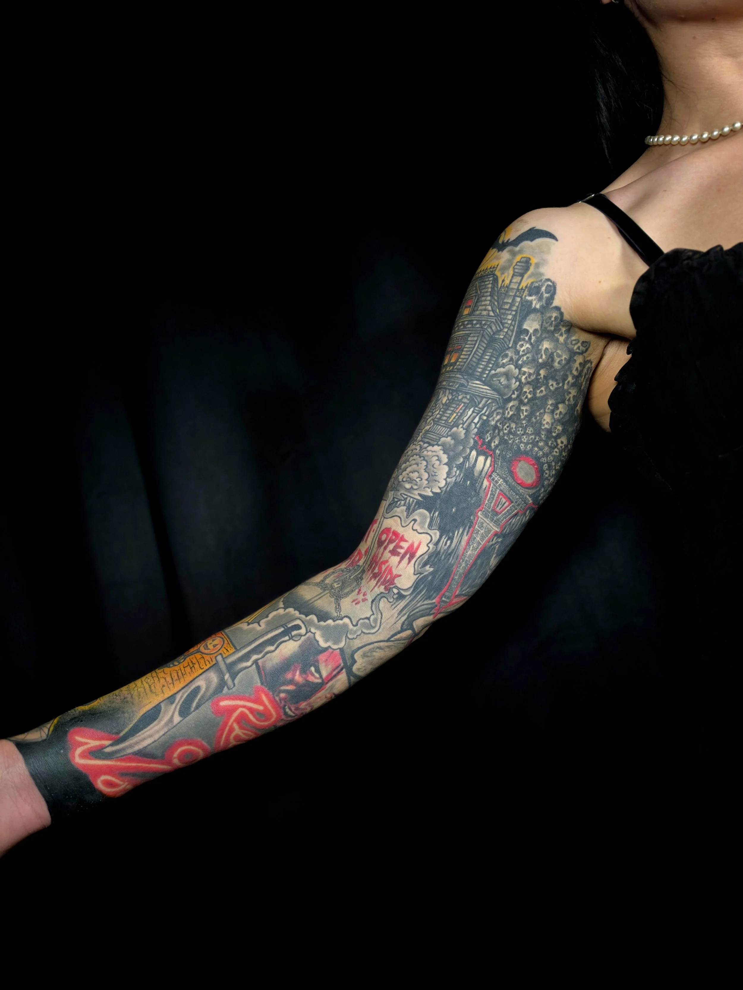 Tattooed arm with colorful artwork, including buildings, flames, and the word 'OPEN' on the forearm. The person is wearing a black off-shoulder top, a pearl necklace, and a black bracelet.