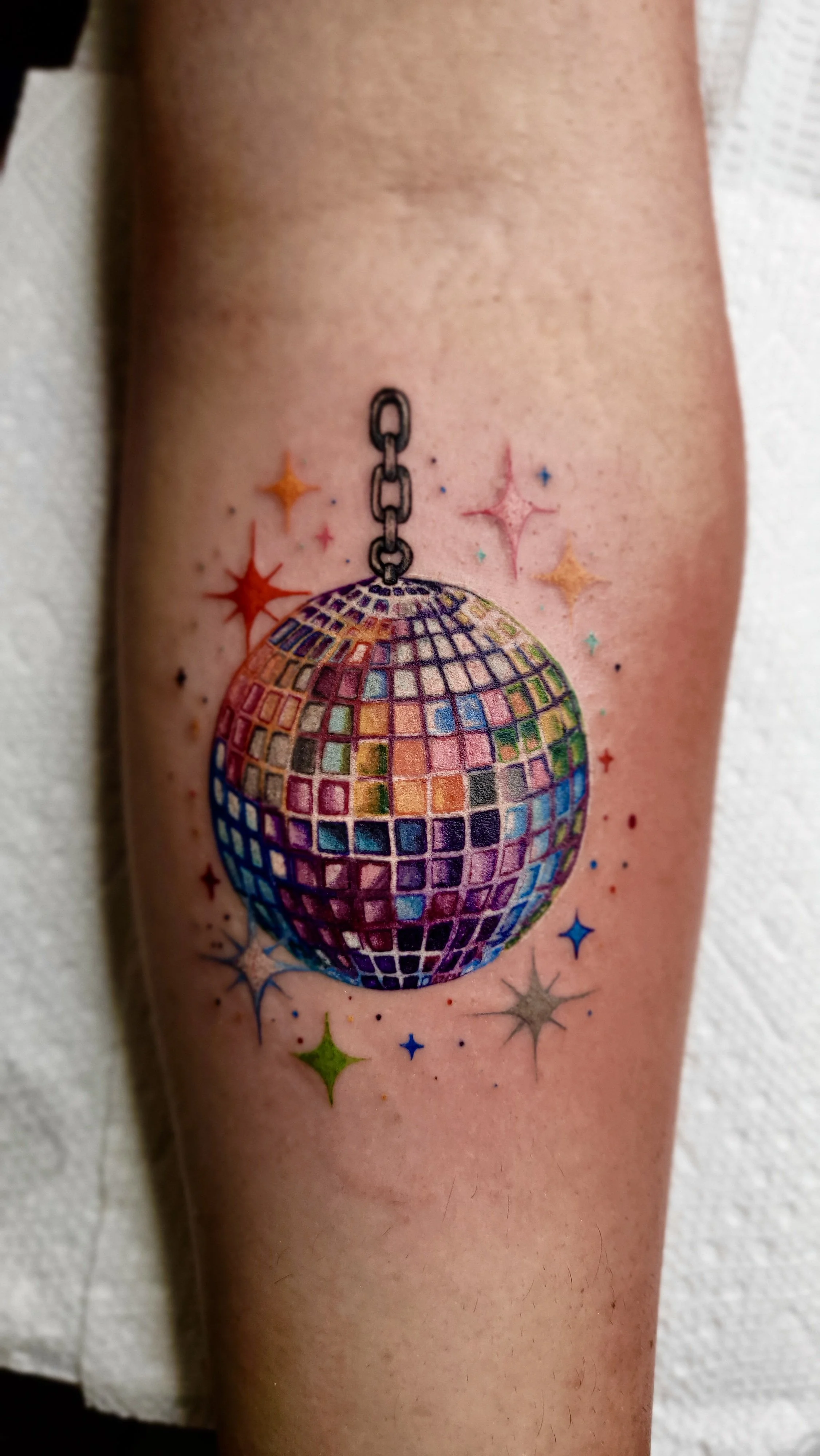 Tattoo of a disco ball hanging from a chain, surrounded by colorful stars and dots.